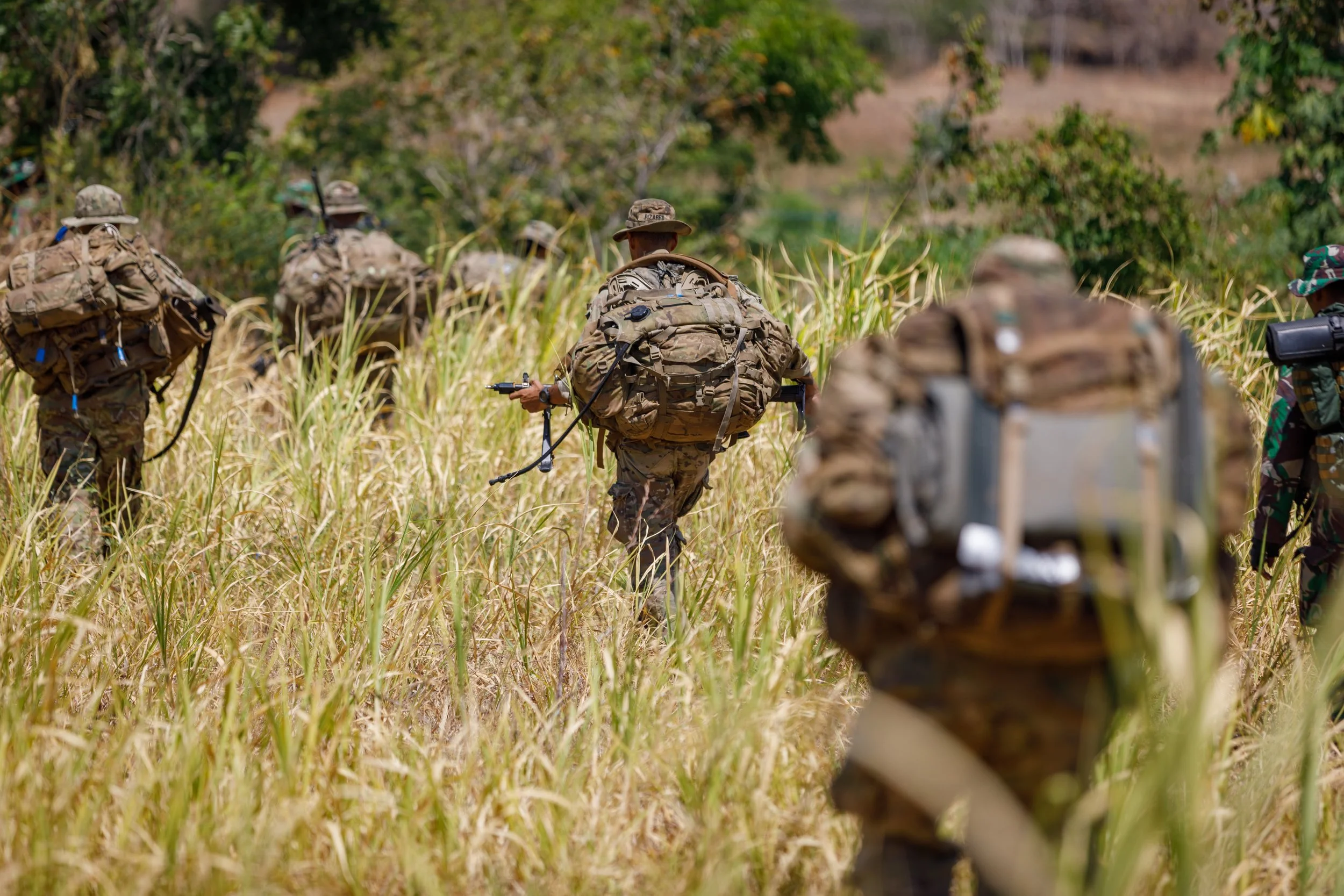 Military soldiers walking through tall grass in a field during daytime, carrying backpacks and gear.