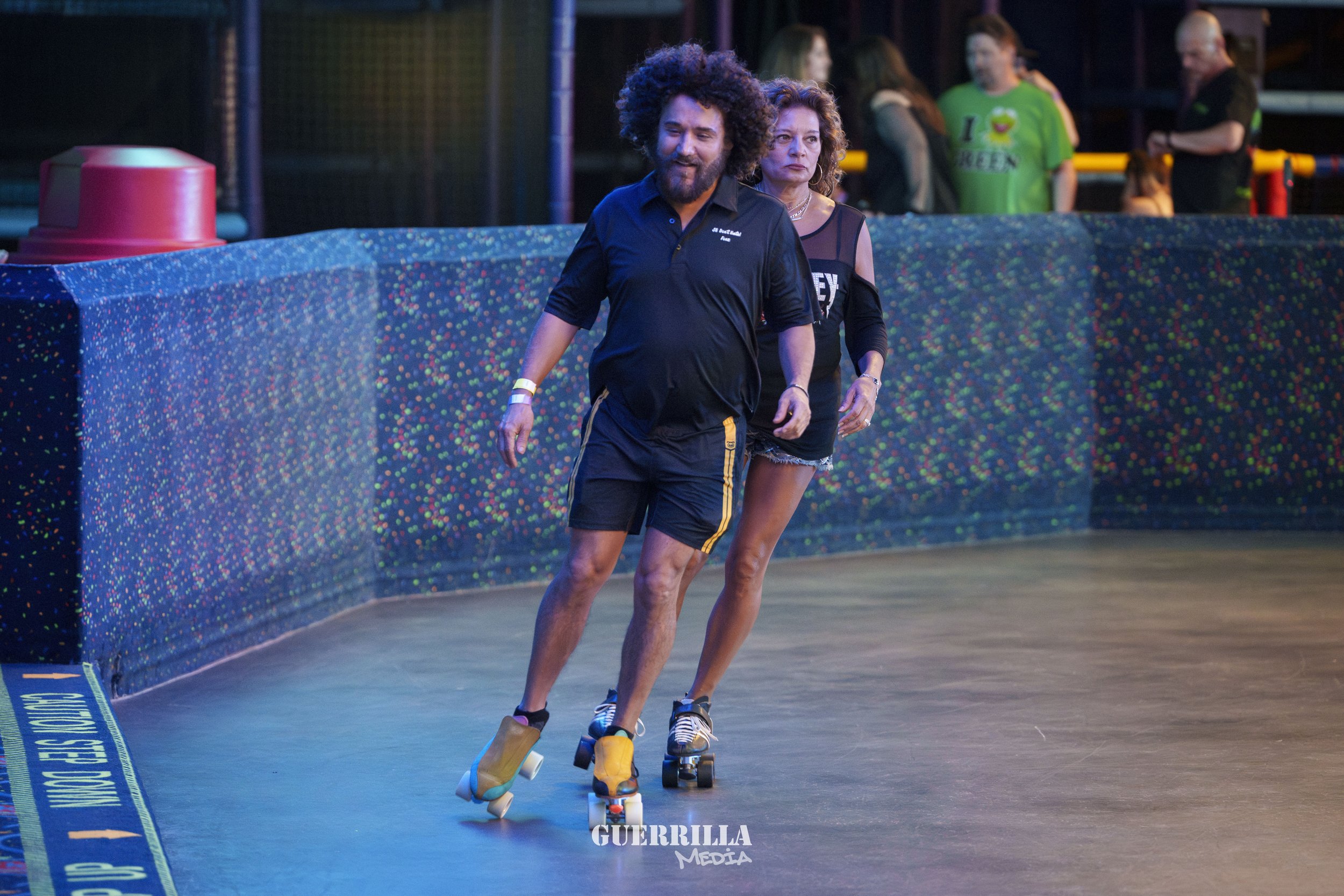 Two people roller skating on an indoor rink, with a colorful dotted barrier in the background and other skaters in the distance.