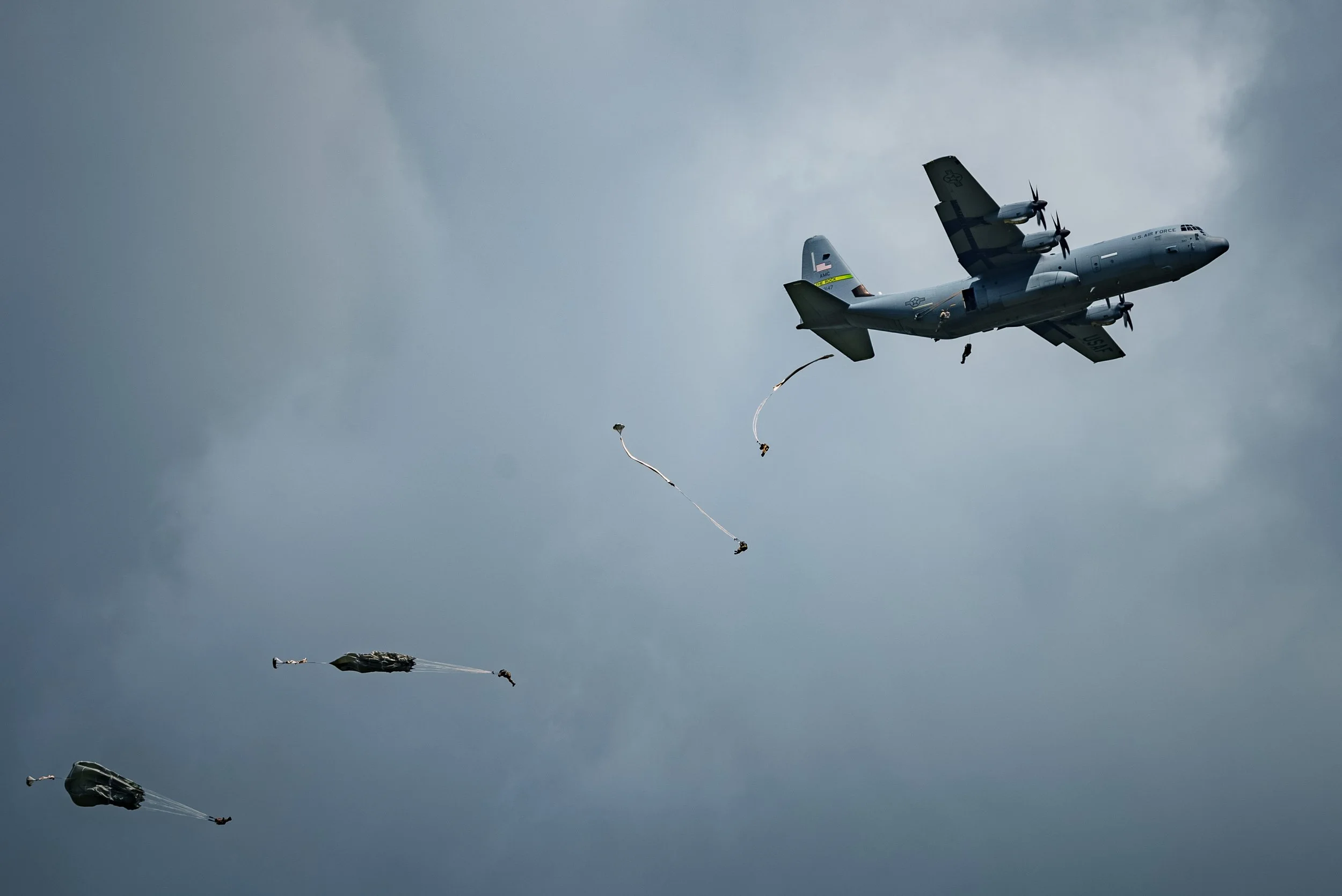 A military cargo plane dropping paratroopers from the sky with dark clouds in the background.