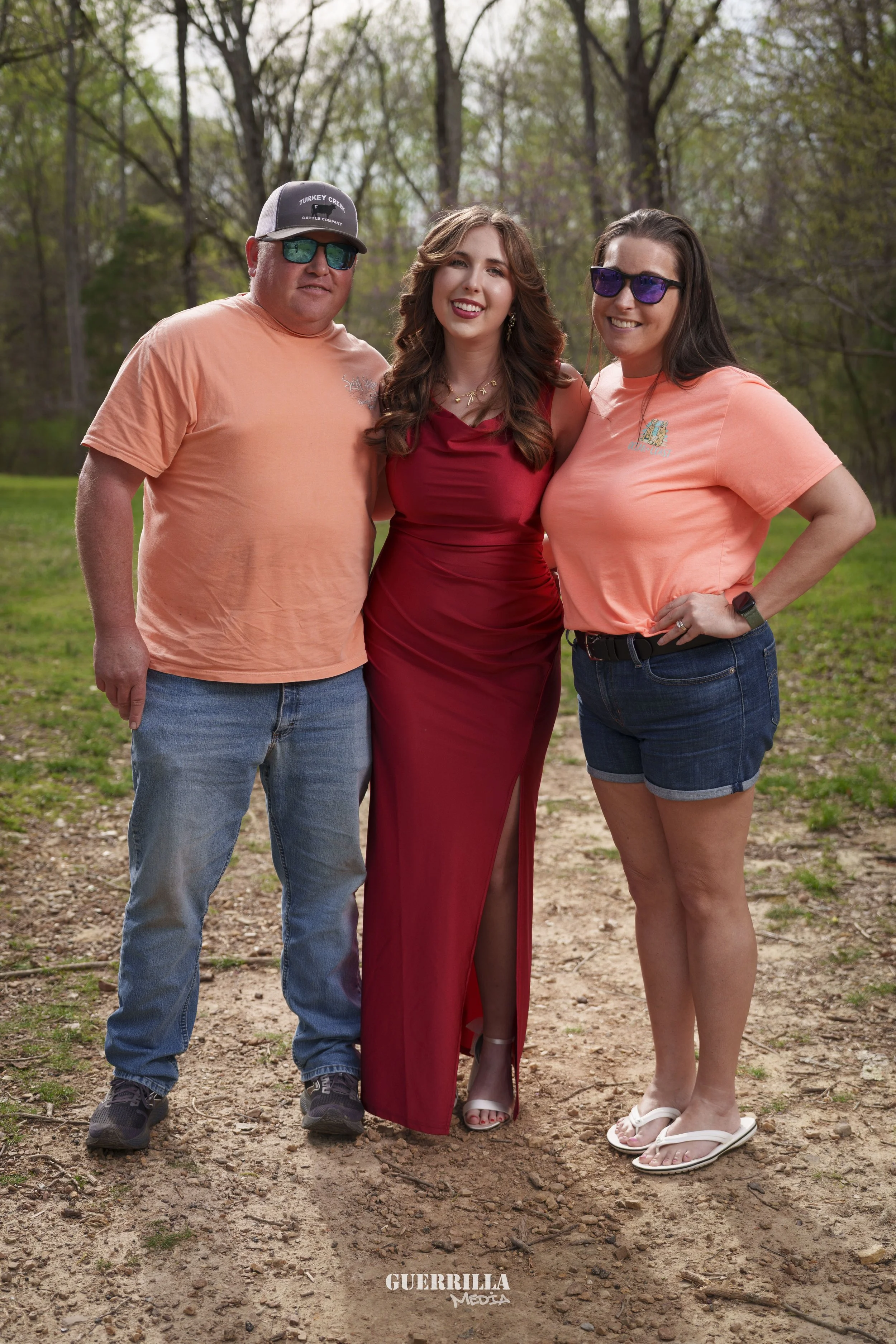 Three people standing outdoors on a dirt path with trees in the background. The man on the left wears a salmon t-shirt, jeans, athletic shoes, sunglasses, and a cap. The woman in the middle wears a red dress and heels. The woman on the right wears a 