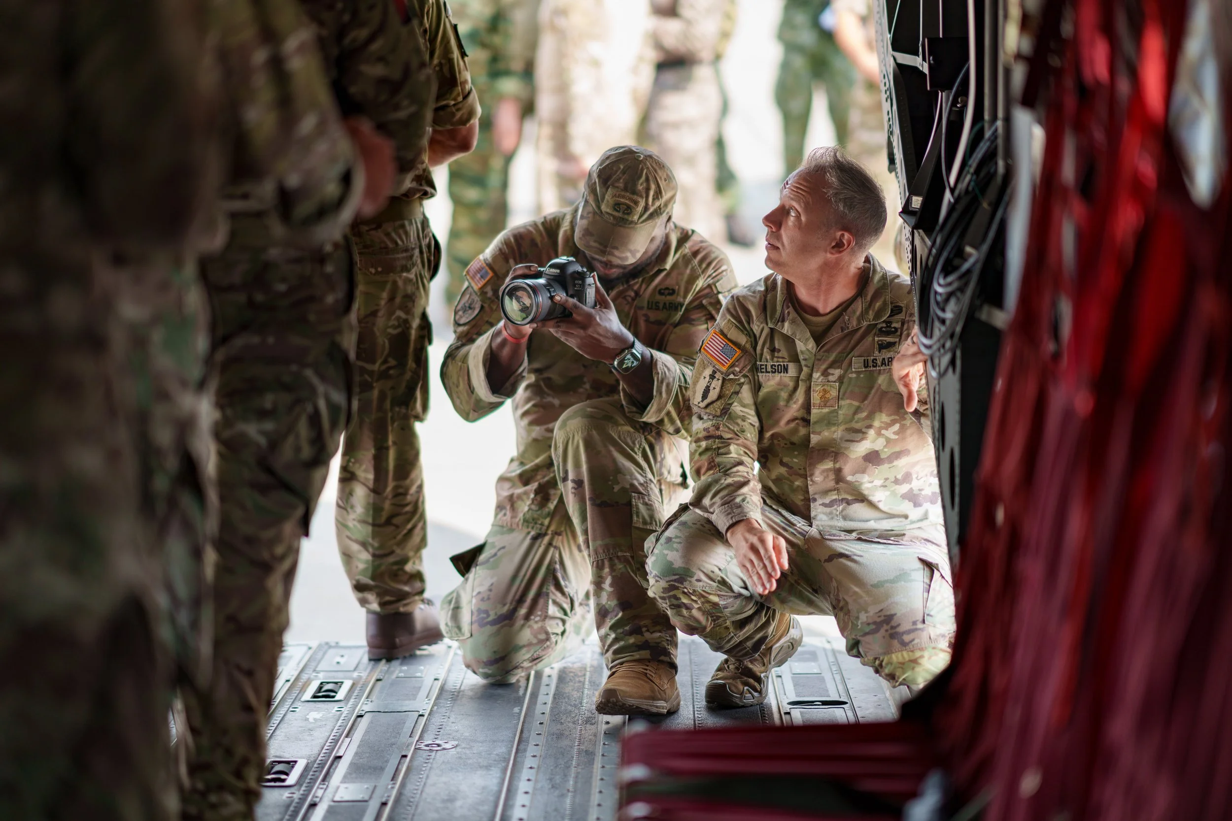 Two soldiers in camouflage uniform kneeling and sitting inside a military aircraft, with one soldier showing a camera to others.