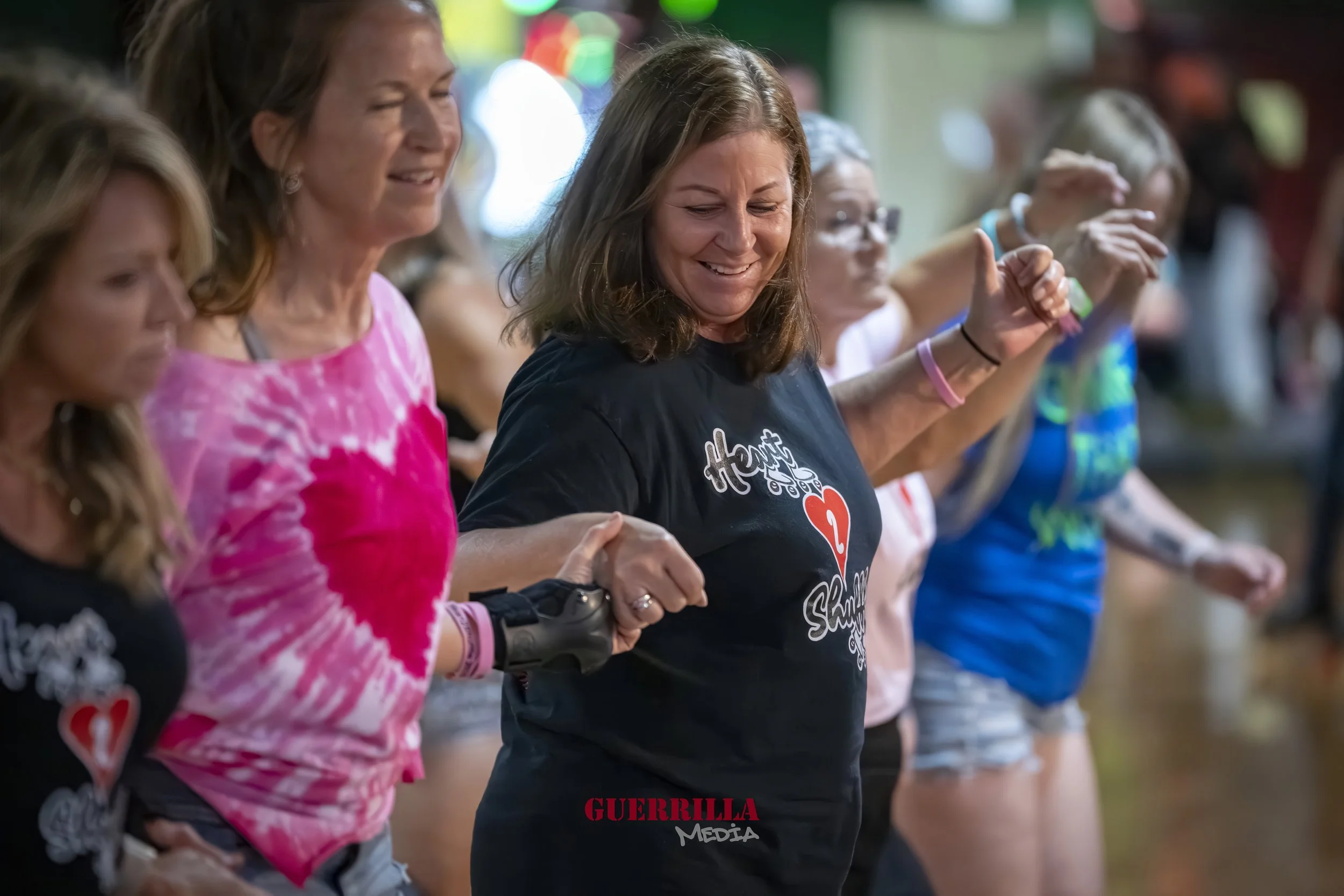 A group of women holding hands in a line, participating in a heartfelt or celebratory event, smiling, with some wearing black T-shirts with a red heart and text.