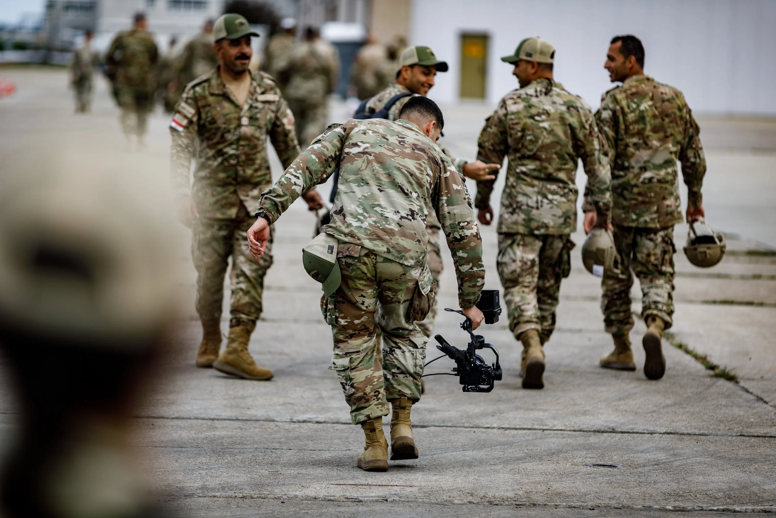 Group of soldiers in camouflage uniforms walking together on a concrete ground, some carrying helmets, one using a camera.
