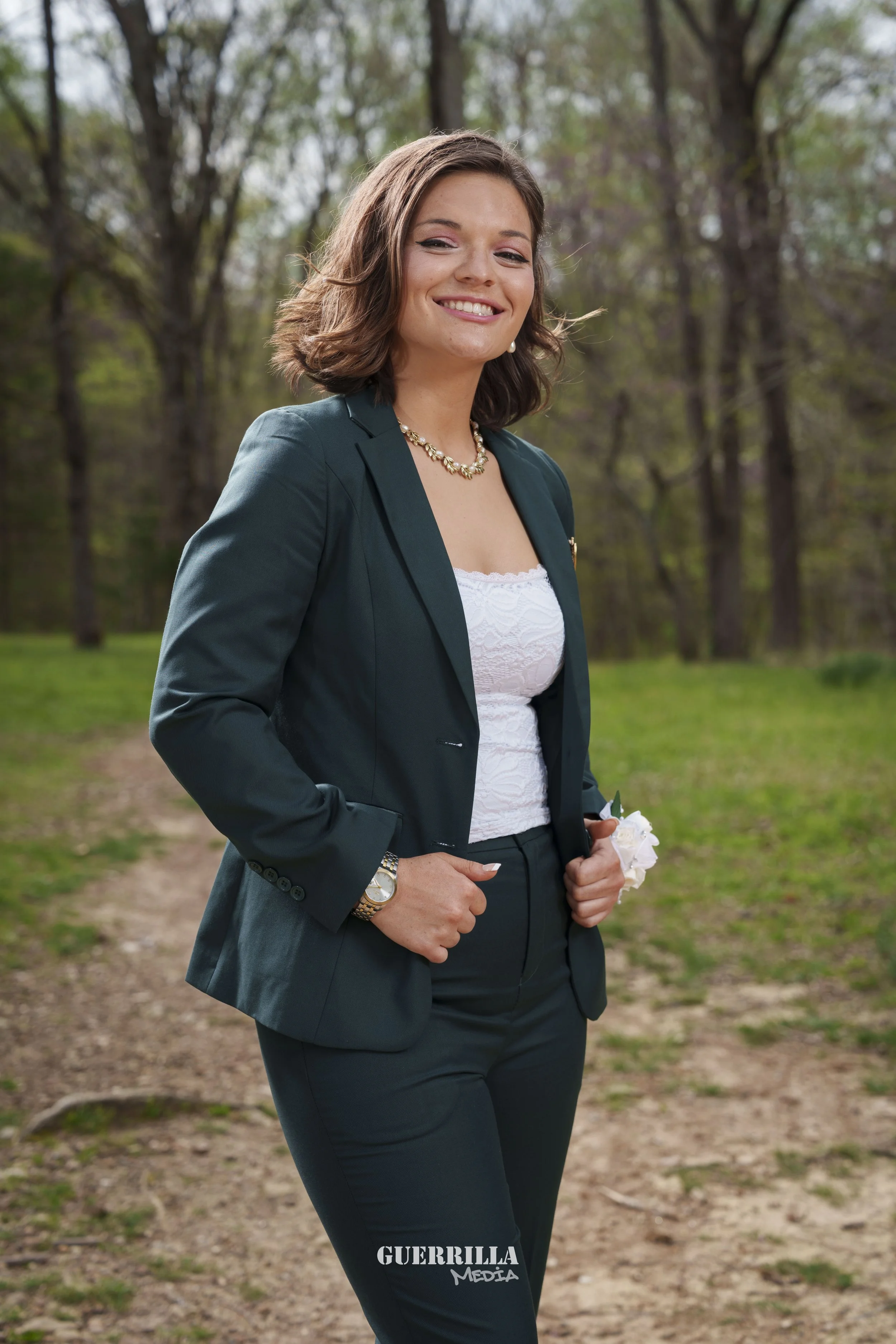 A smiling woman in business attire standing outdoors on a dirt path with trees and green foliage in the background.