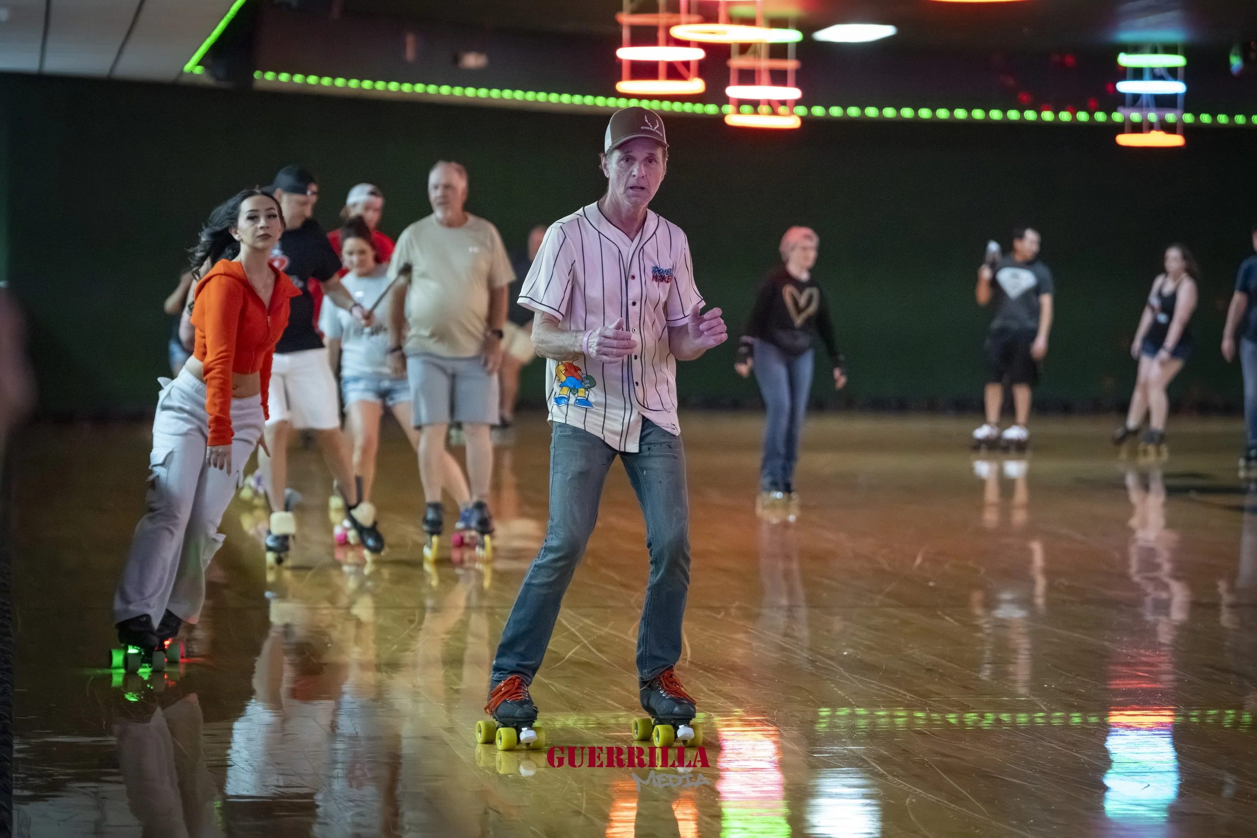 People roller skating at an indoor rink with colorful neon lights overhead, and one person in the foreground wearing a baseball jersey and jeans.