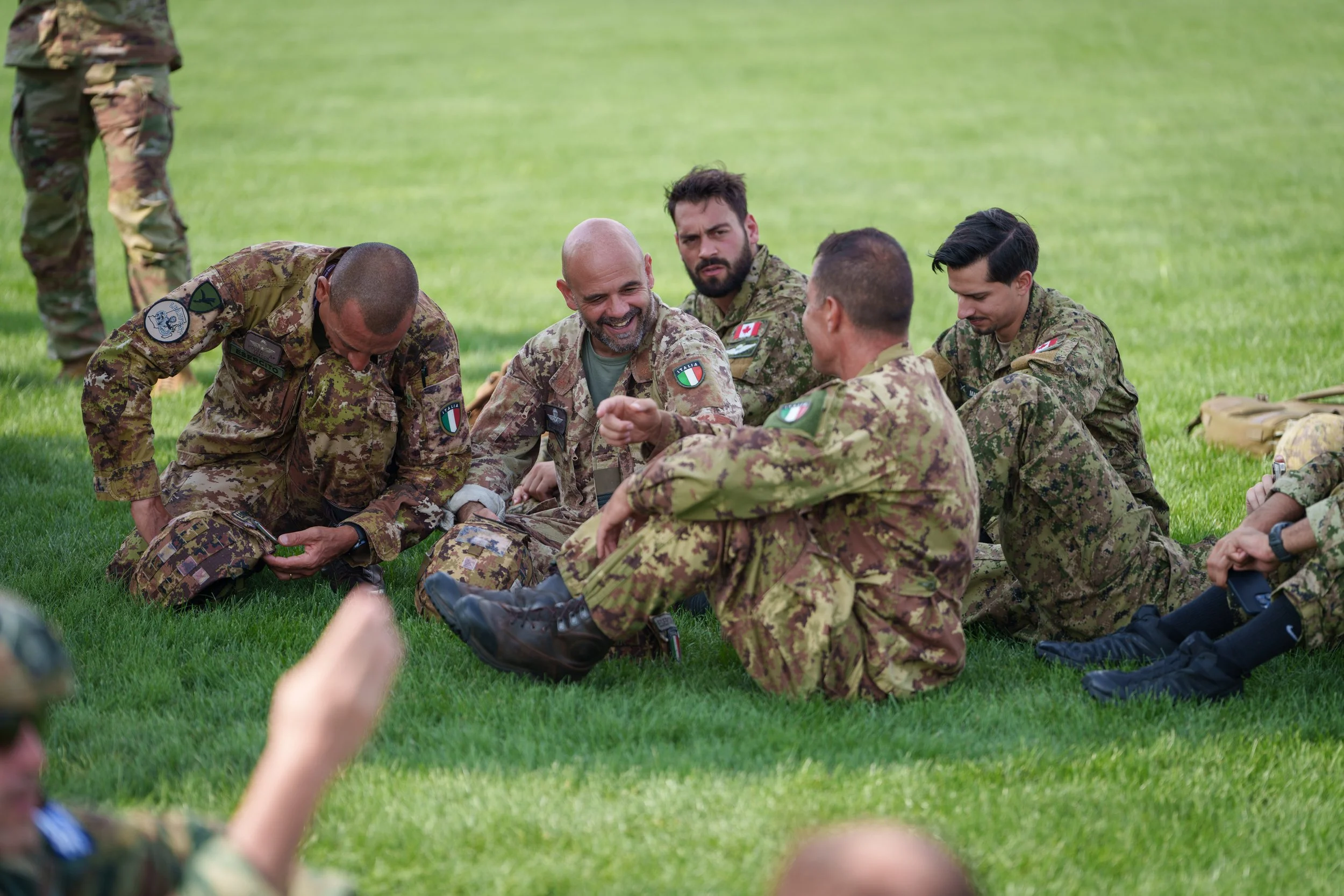Group of soldiers in camouflage uniforms sitting and kneeling on grass, smiling and engaging in conversation outdoors.