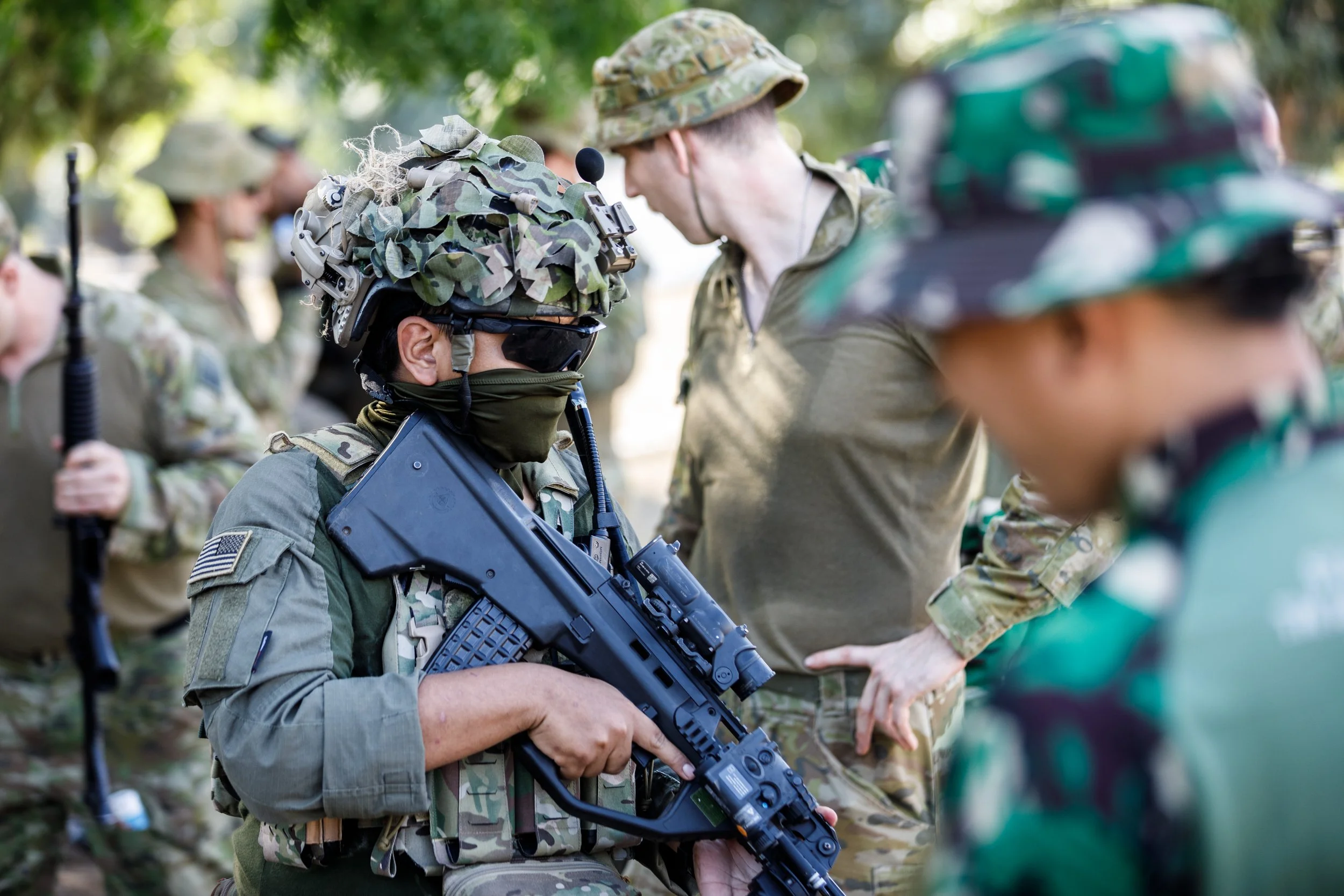 Military soldiers in uniform during a training exercise, with one soldier kneeling and holding a firearm, surrounded by colleagues with serious expressions.