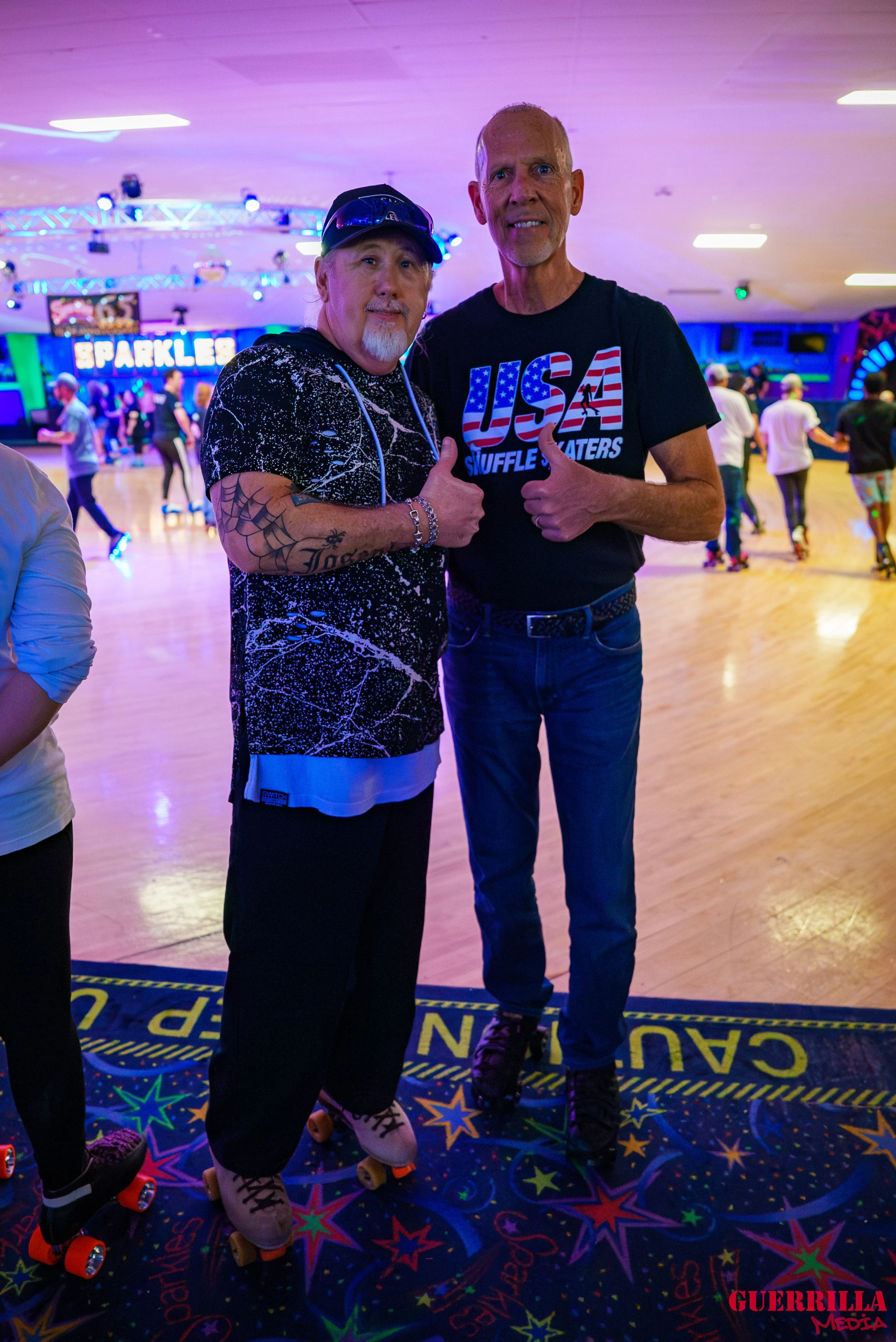 Two men are standing on roller skates in a roller skating rink, posing for the photo with thumbs up. The man on the left has a tattoo on his arm and is wearing a black and white patterned shirt with black pants. The man on the right is wearing a T-sh