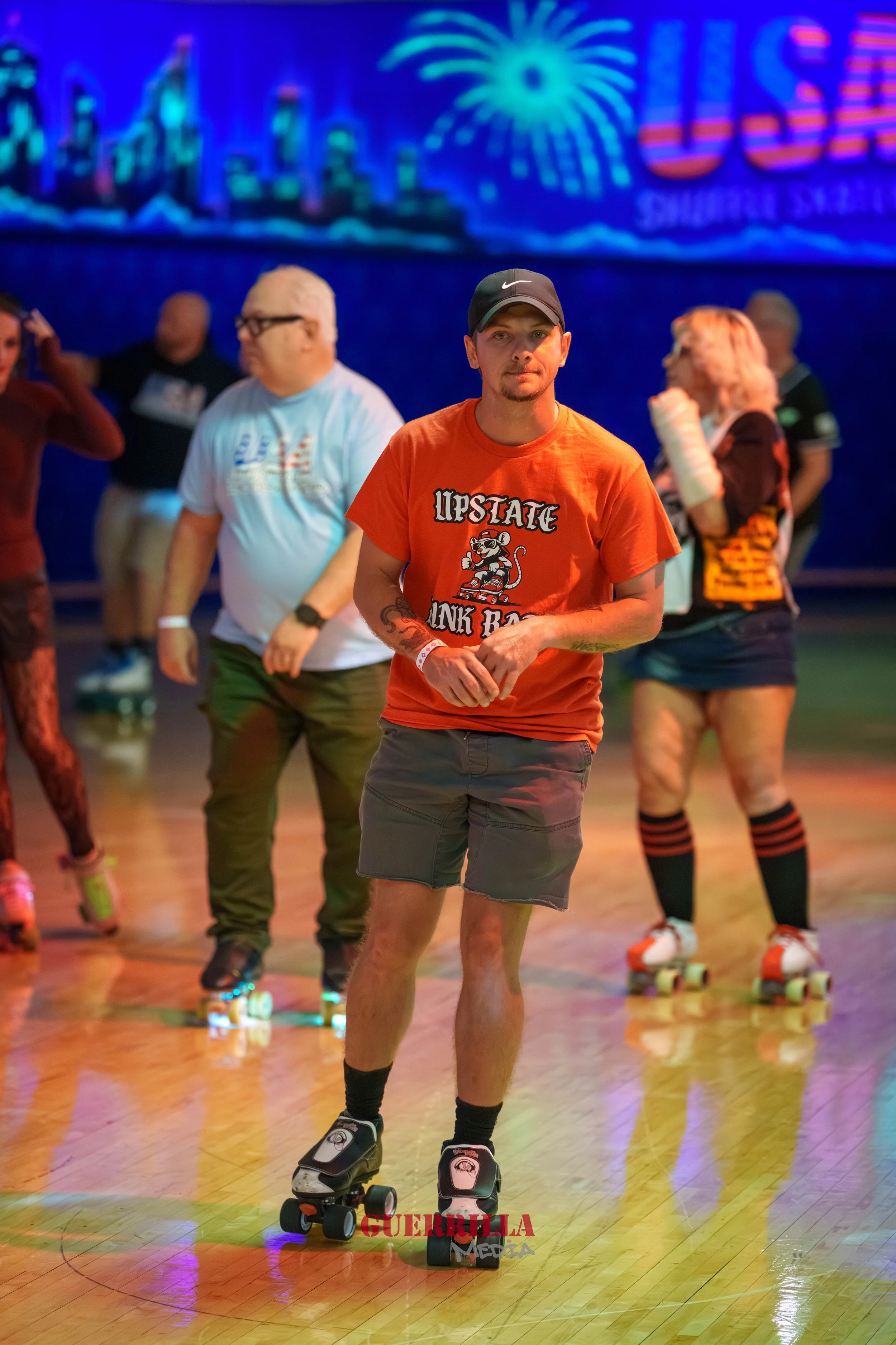 People roller skating indoors, with a younger man in the foreground wearing a black cap and orange T-shirt, with others in the background on a wooden floor, and a cityscape and fireworks display on a large digital screen behind them.