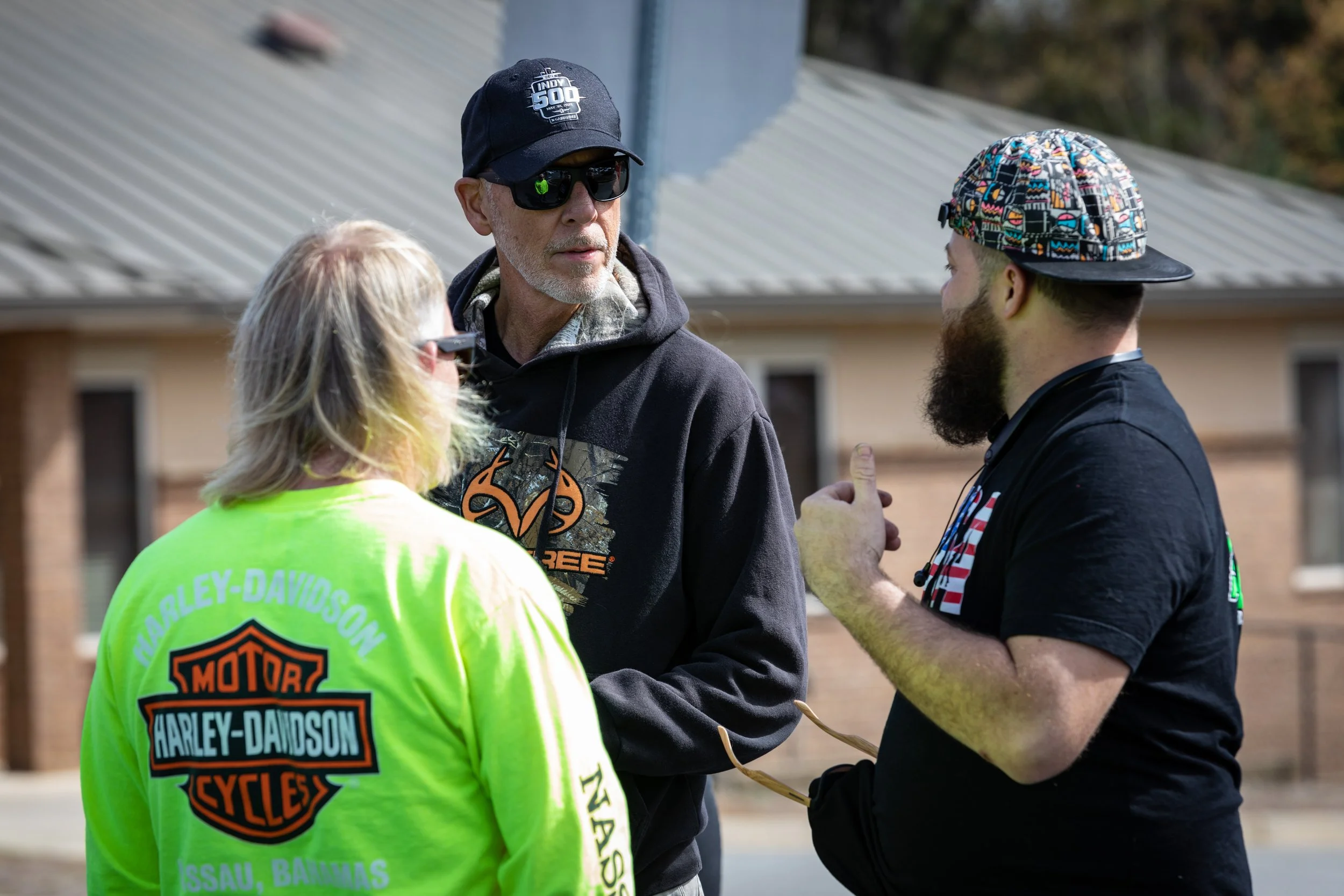 Three people having a conversation outdoors, one wearing a bright yellow Harley-Davidson jacket, another wearing a black hoodie with a logo, and the third in a black shirt with a colorful cap, with buildings in the background.