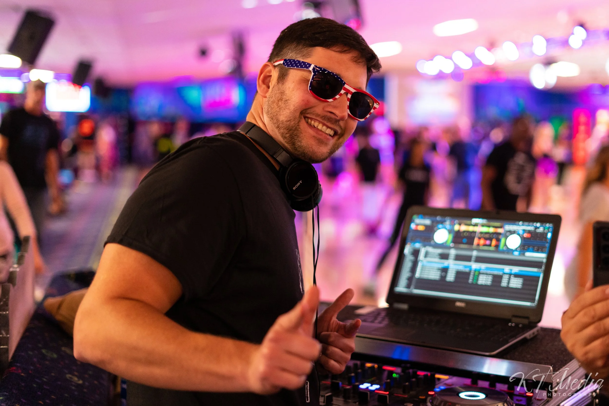 A young man with short dark hair, wearing patriotic American flag sunglasses, smiling and giving a thumbs up at a nightclub or party, standing in front of a DJ setup with a laptop and turntables, with colorful blurred lights and people in the backgro