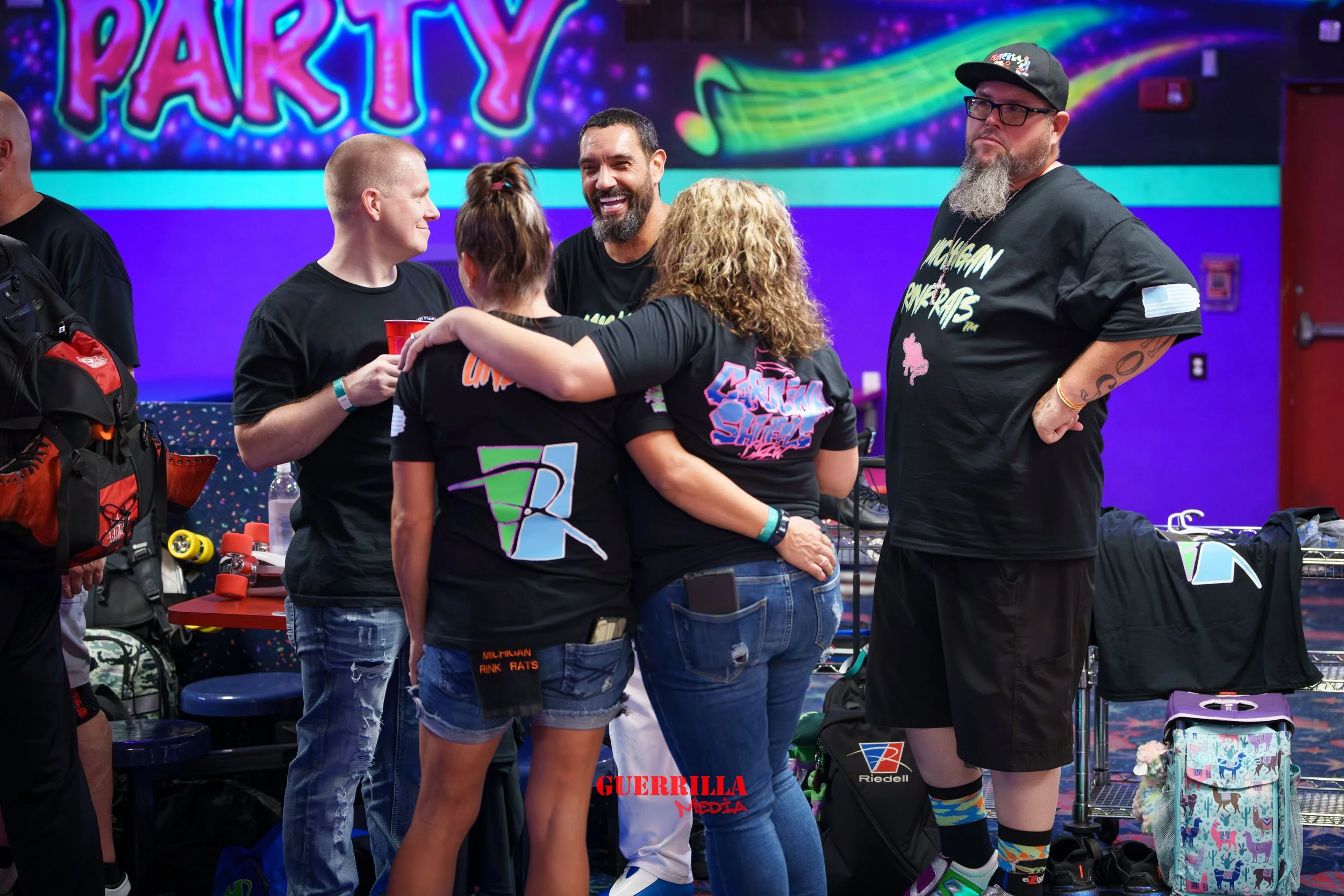 Group of five people at a party in an arcade or entertainment venue with colorful lighting and neon signs, including a large 'PARTY' sign in the background. They are wearing casual clothes, some with graphic T-shirts, and are engaging in conversation