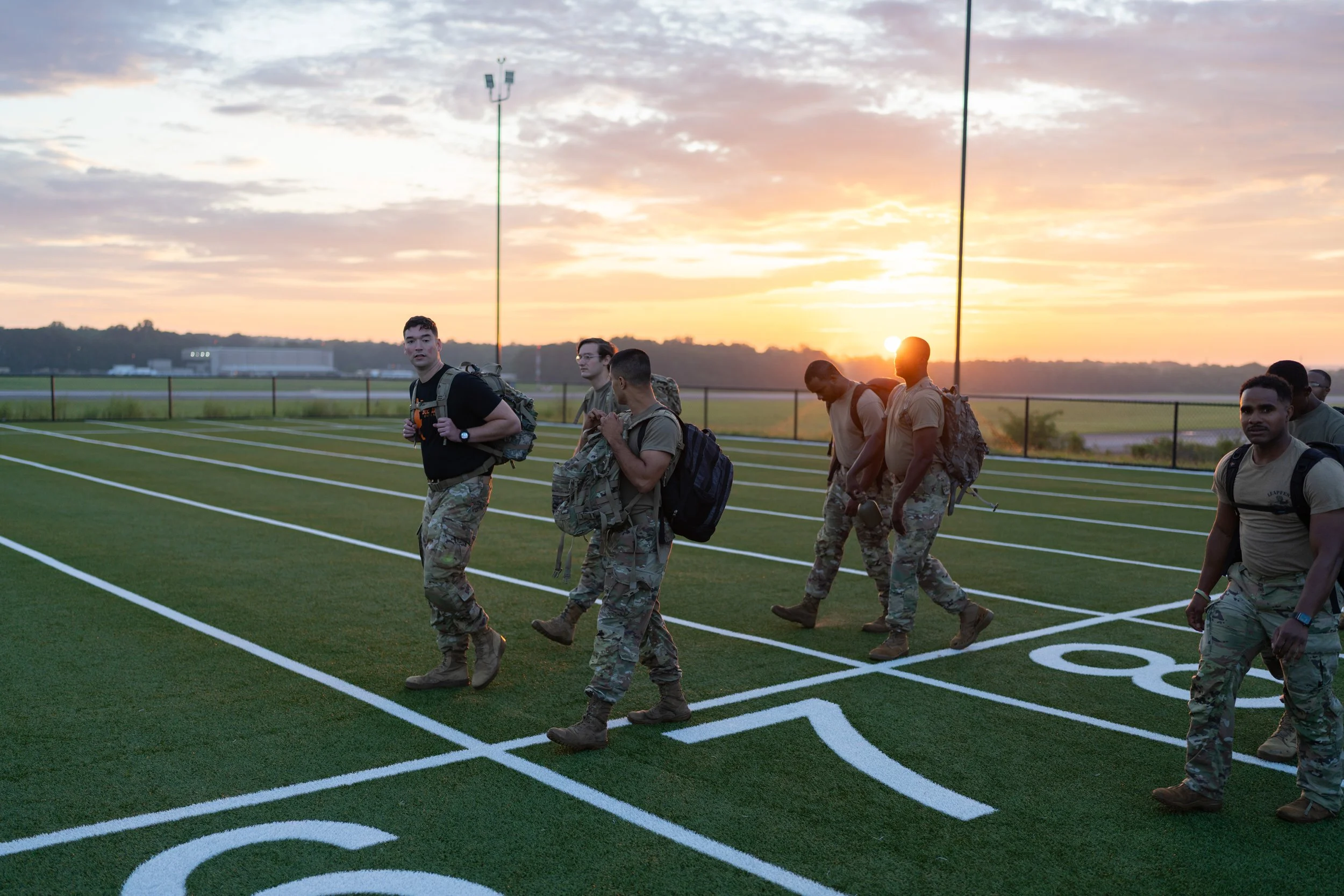 Group of soldiers walking on a running track at sunset, wearing military uniforms and backpacks.