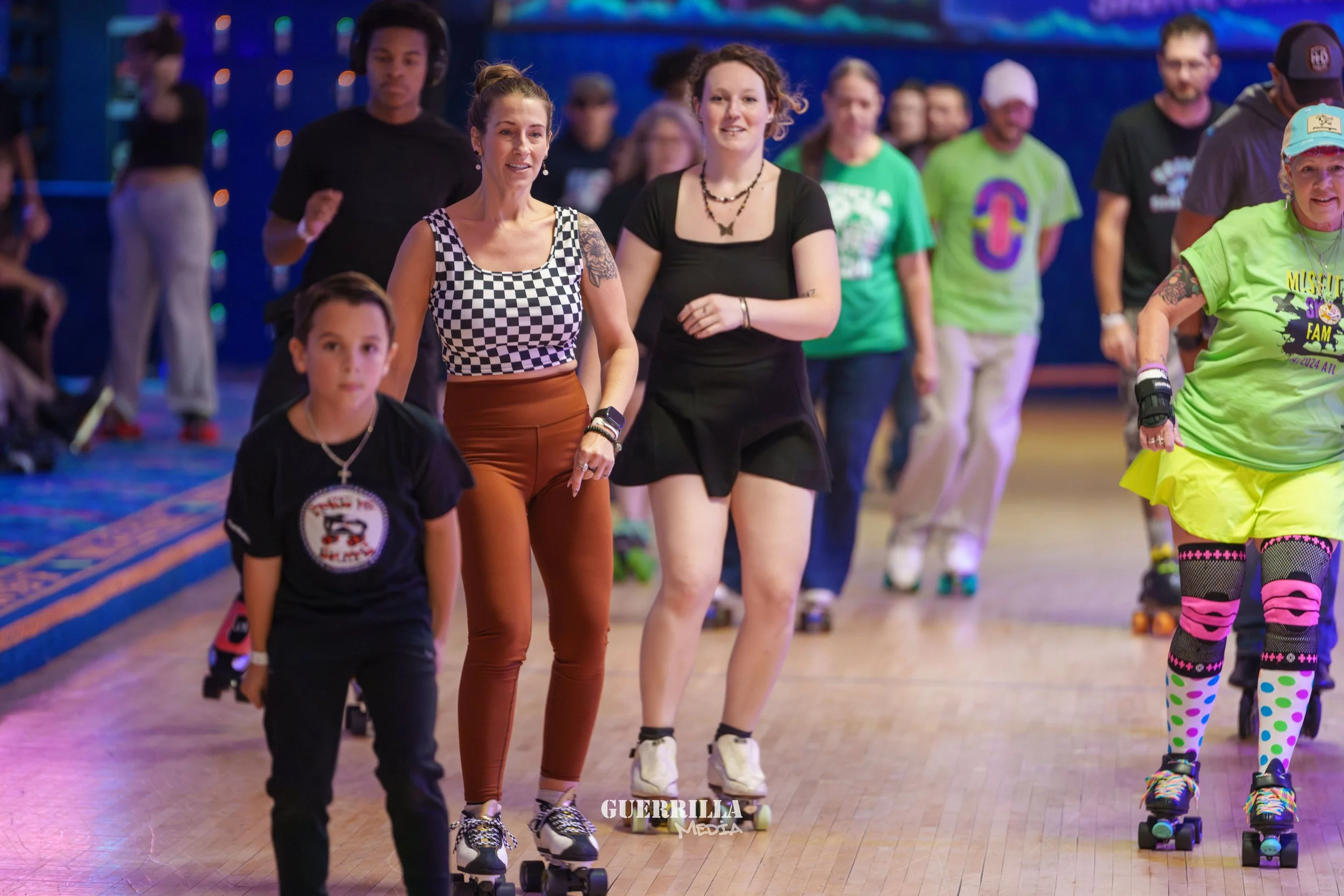 Group of people roller skating indoors, including a young girl in black, two women in colorful outfits, and others in the background on a wooden rink.