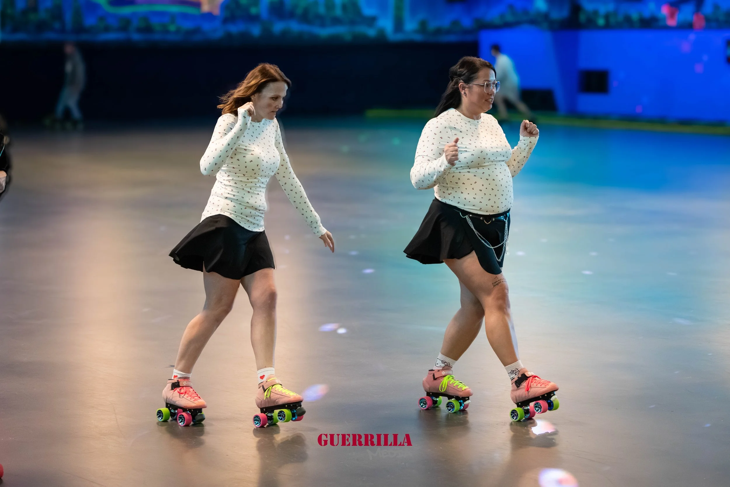 Two women roller skating indoors, wearing matching white polka dot sweaters and black skirts, with one cheerfully dancing while the other is looking down, in a brightly lit space.