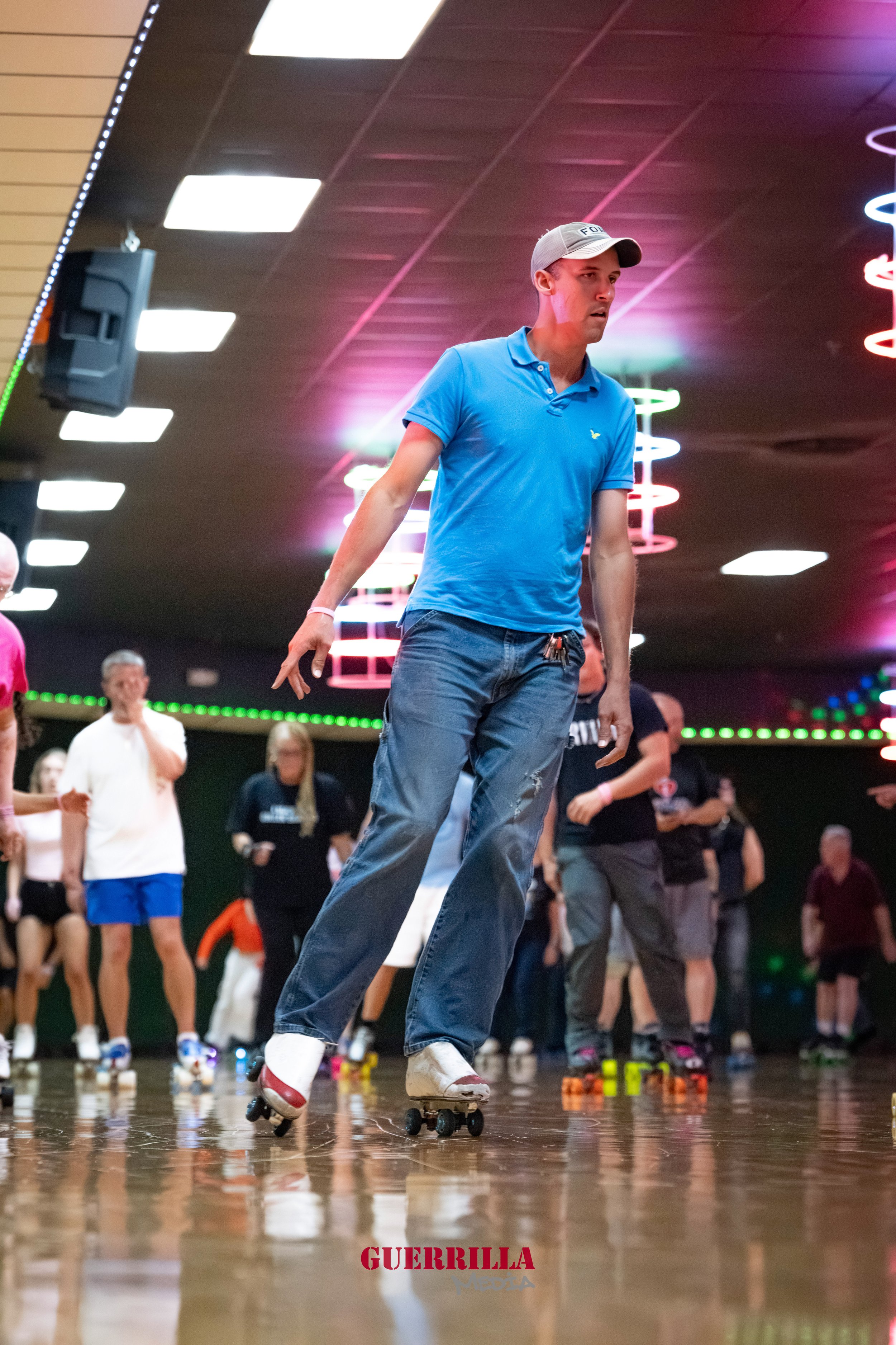 A man wearing a gray cap and blue polo shirt roller skating in an indoor roller rink with an audience in the background.