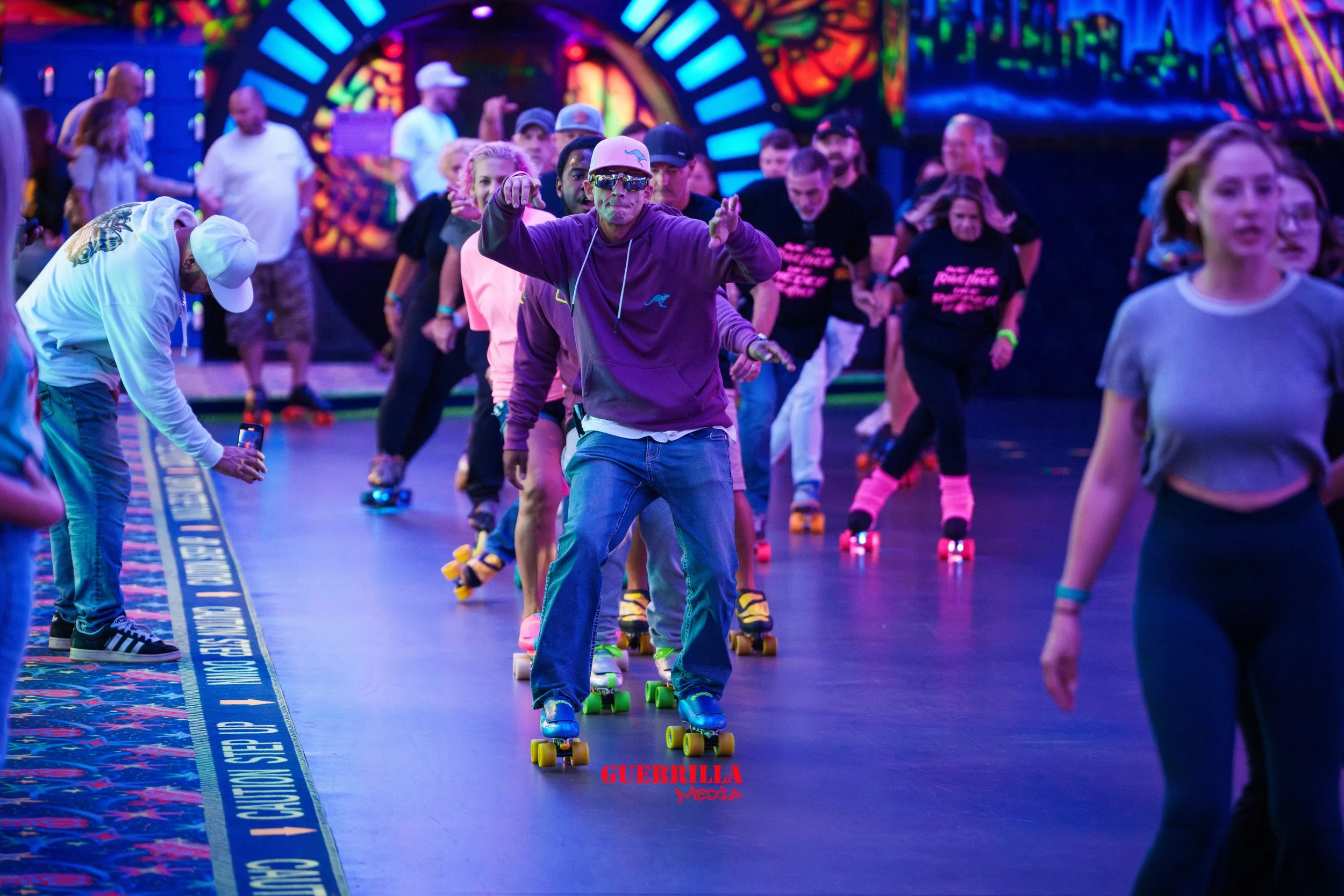 Group of people roller skating indoors with vibrant, colorful lights and decorations in the background. Some individuals are taking photos, while others are skating and enjoying the activity.