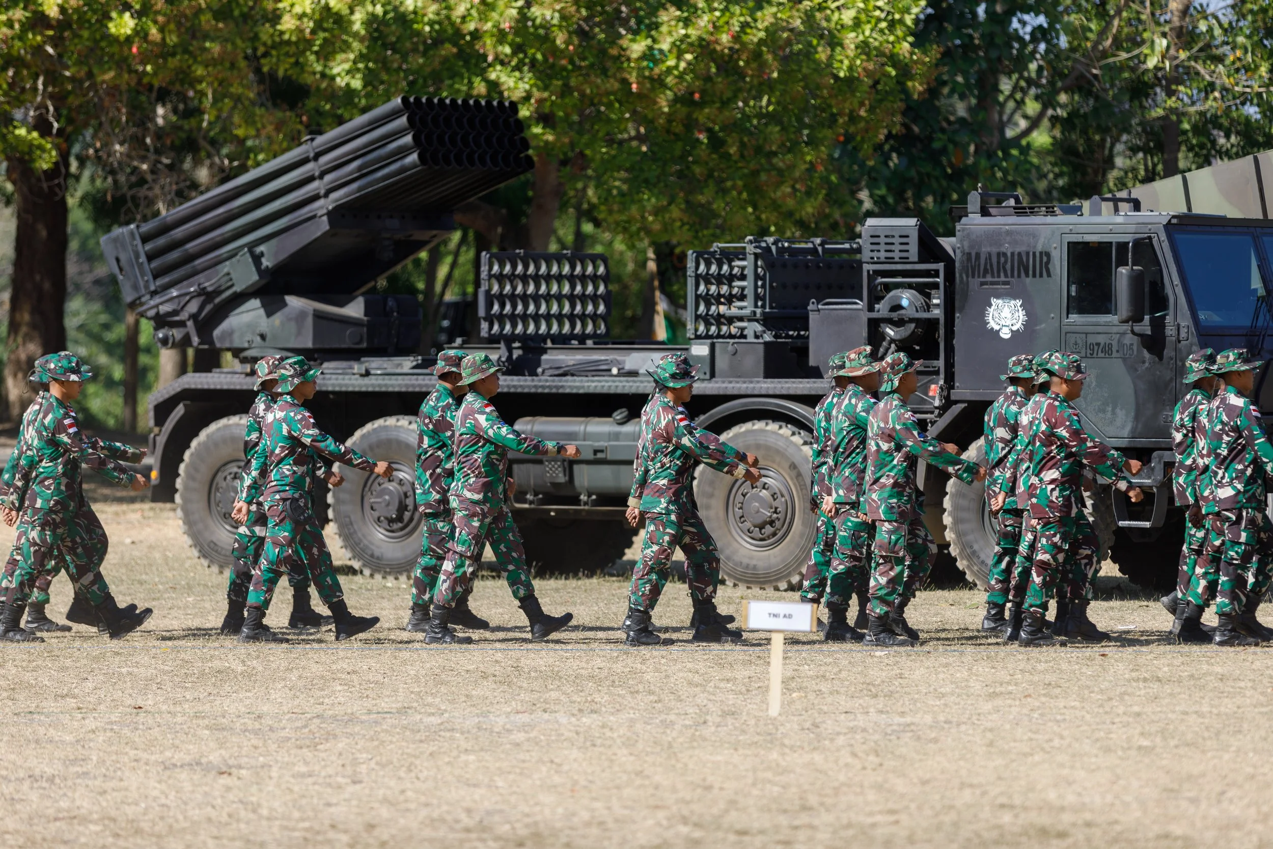 A group of soldiers in camouflage uniforms walking in a line near a military vehicle with a rocket launcher on top, on a grassy field with trees in the background.