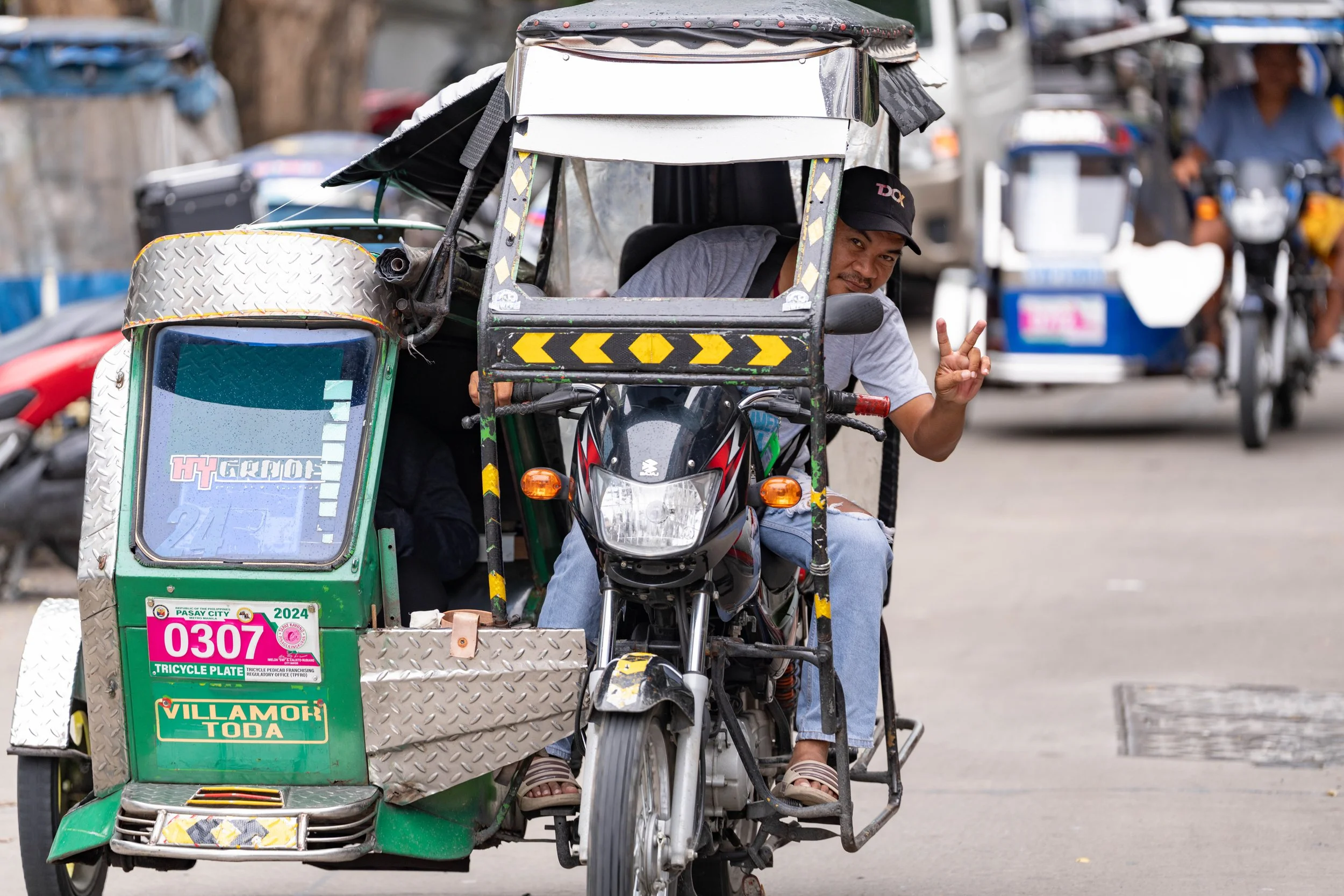 A man in a baseball cap riding a motorcycle that has a sidecar with a passenger inside. The man is making a peace sign with his fingers and looking at the camera.