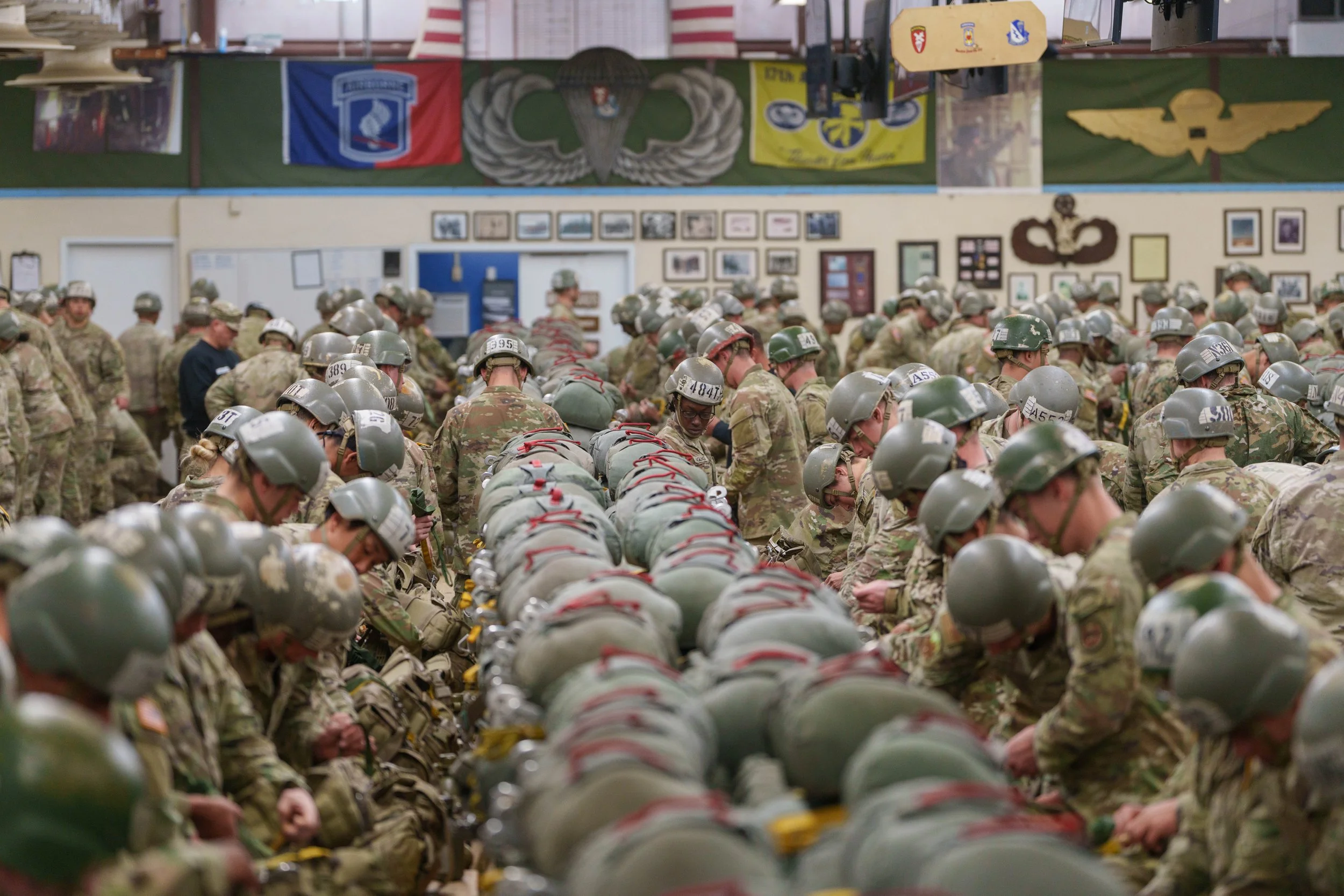 A large group of soldiers in camouflage uniforms and helmets, kneeling with bowed heads, inside a room decorated with military banners, flags, and framed pictures.