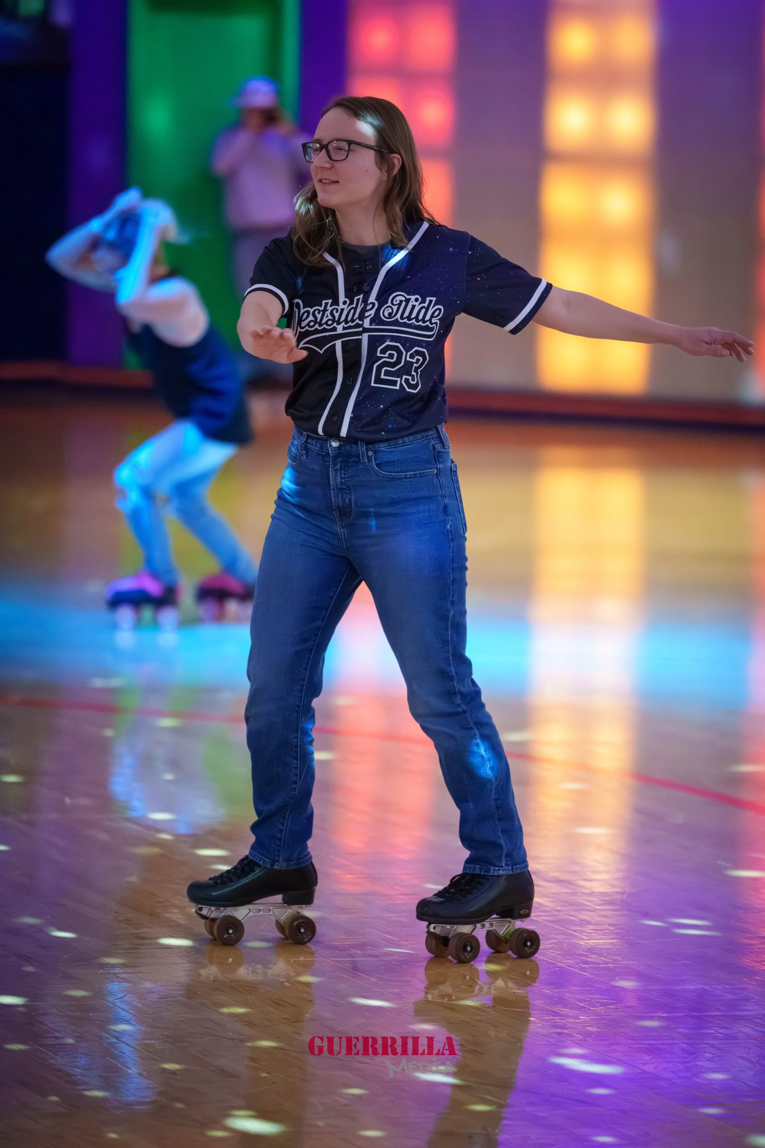 A woman roller skating indoors at a roller rink, with a person in the background wearing roller skates and a hat. The woman has glasses, long hair, and is wearing a black baseball jersey with the number 23, and blue jeans.