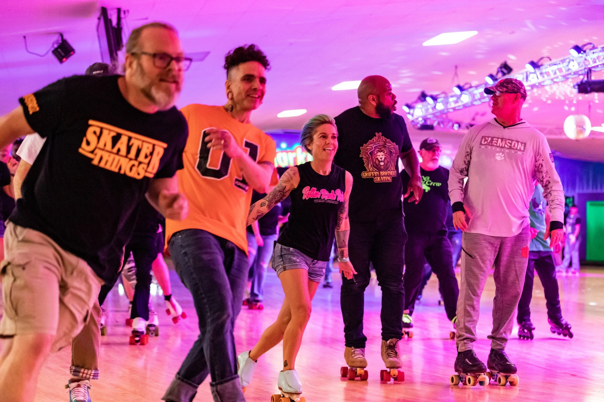 People roller skating in a well-lit indoor rink with pink and purple lighting, smiling and holding hands while skating together.