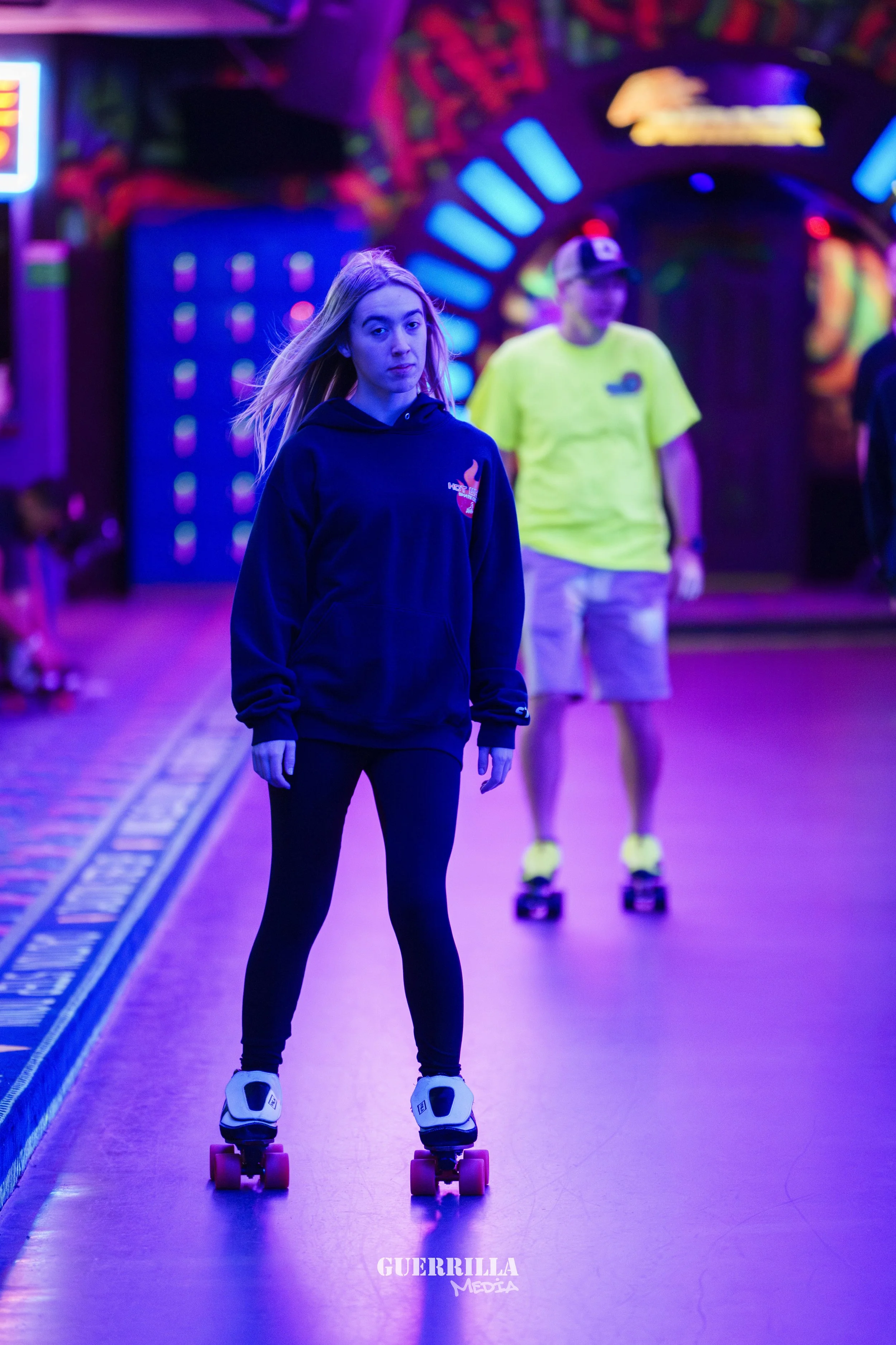 A young woman with long blonde hair skateboarding inside a glow-in-the-dark amusement park or arcade with neon lighting. She is wearing a black hoodie and black leggings. In the background, a man wearing a yellow shirt and cap is also on a skateboard