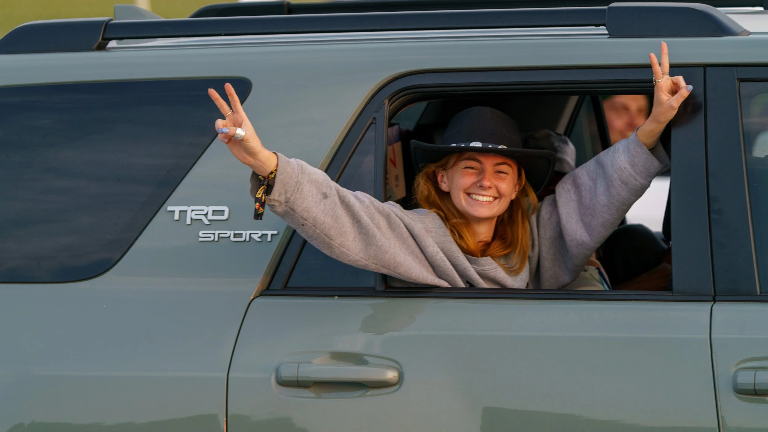 A happy woman with red hair smiling and making peace signs with both hands through the open window of a green SUV, wearing a large black hat and a gray sweatshirt.