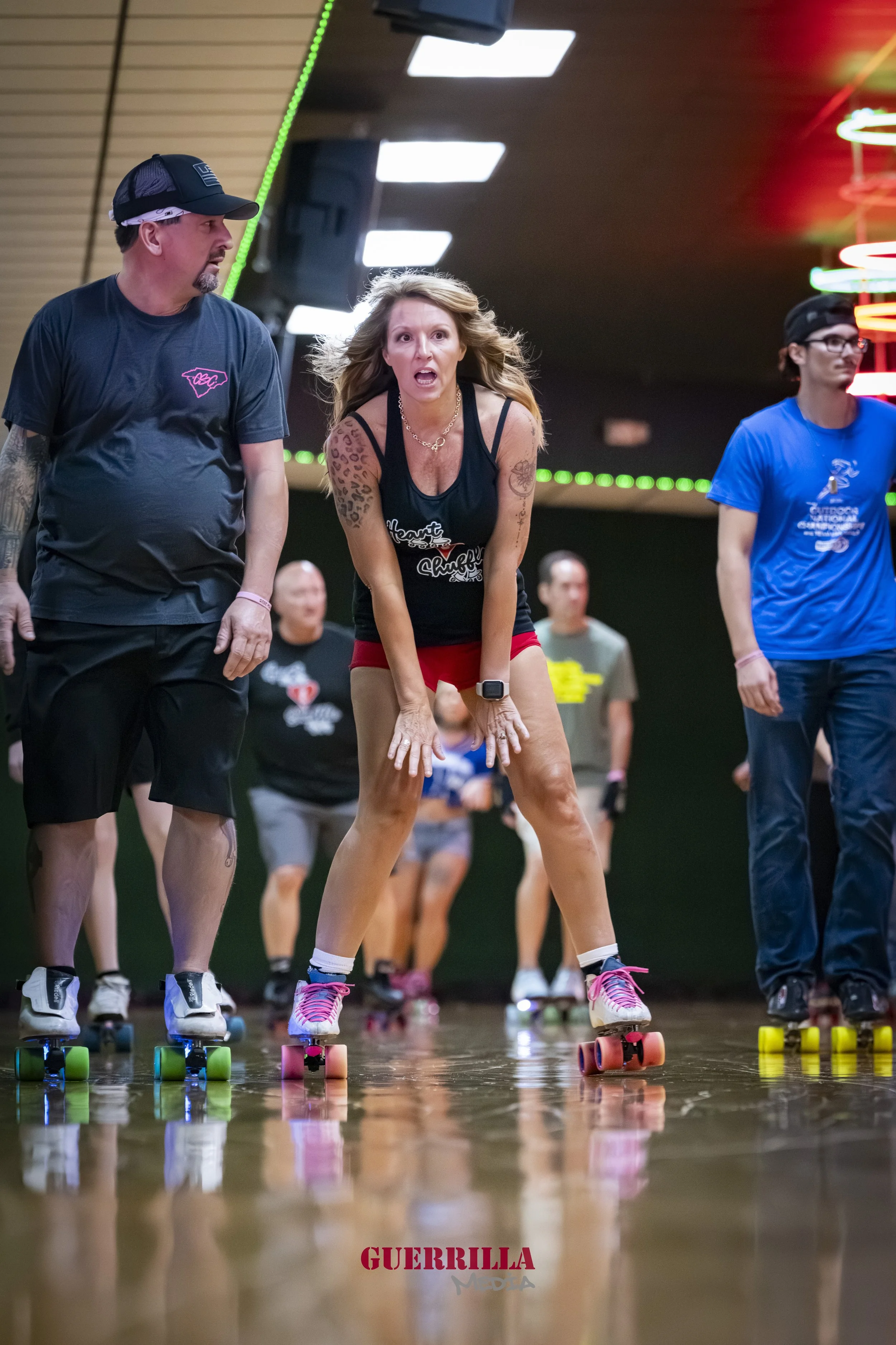 A group of people roller skating indoors. A woman in the foreground looks surprised or excited, wearing a black tank top and red shorts. Several others are visible in the background, all skating on a polished wooden floor under colorful neon lights.