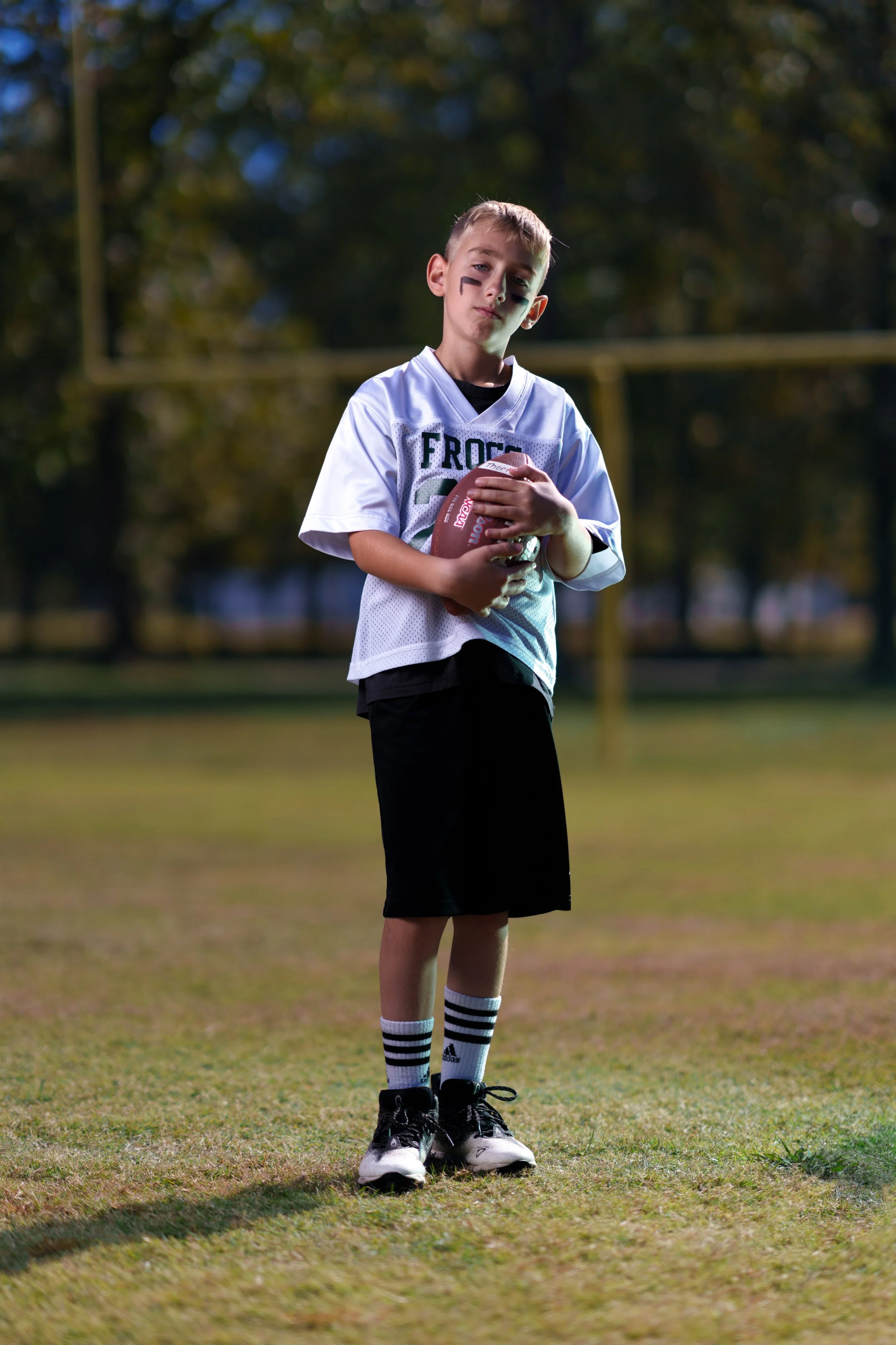 A young boy in American football gear standing on a grassy field holding a football, with a football field goal post and trees in the background.