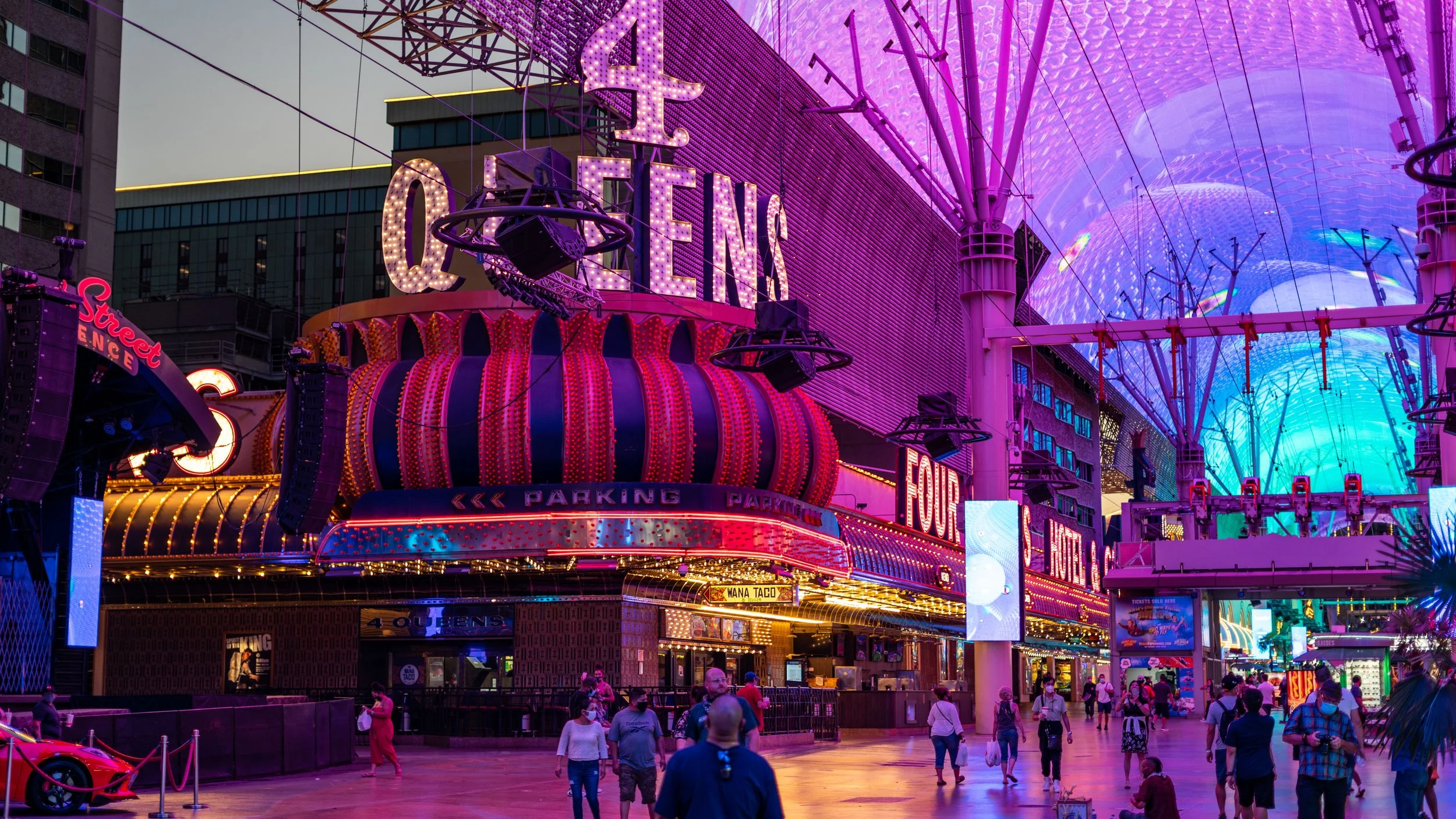 Night view of Las Vegas Fremont Street with bright neon lights, including signs for the Queen's casino, a large pumpkin-shaped light structure, and the neon canopy overhead; people walking in the street.