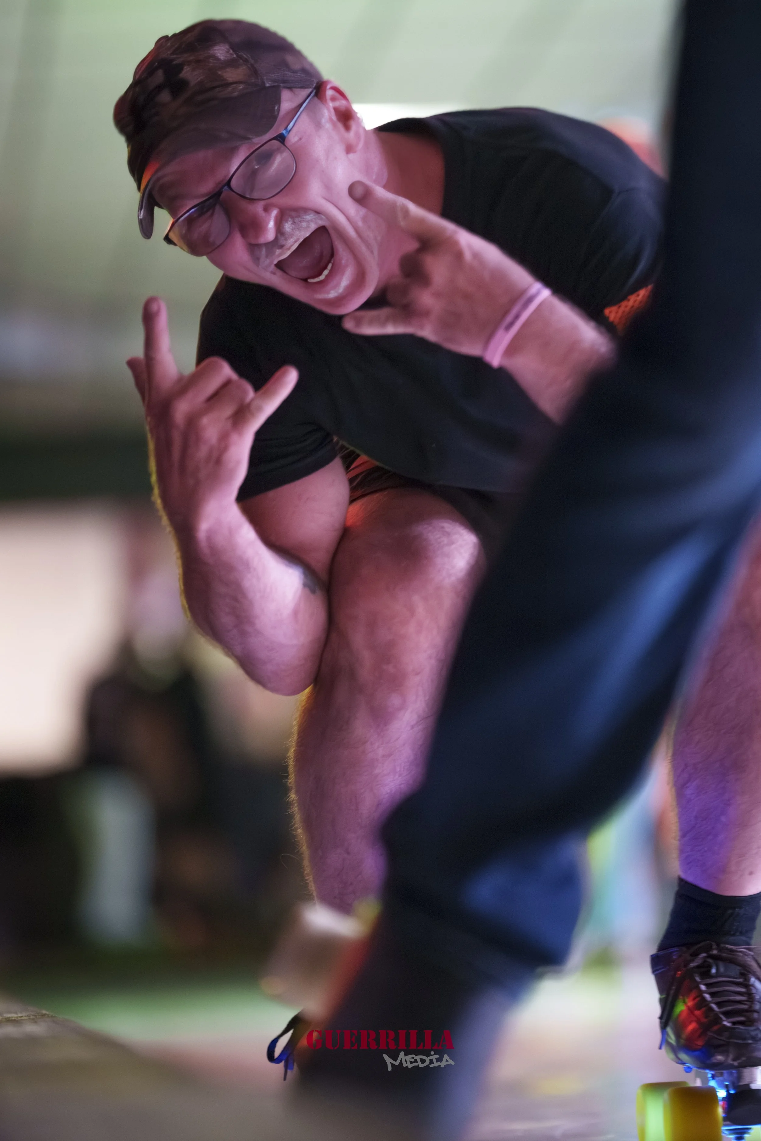 Man in black shirt making a rock and roll hand gesture, smiling and shouting, with curly hair and glasses, at a roller derby event.