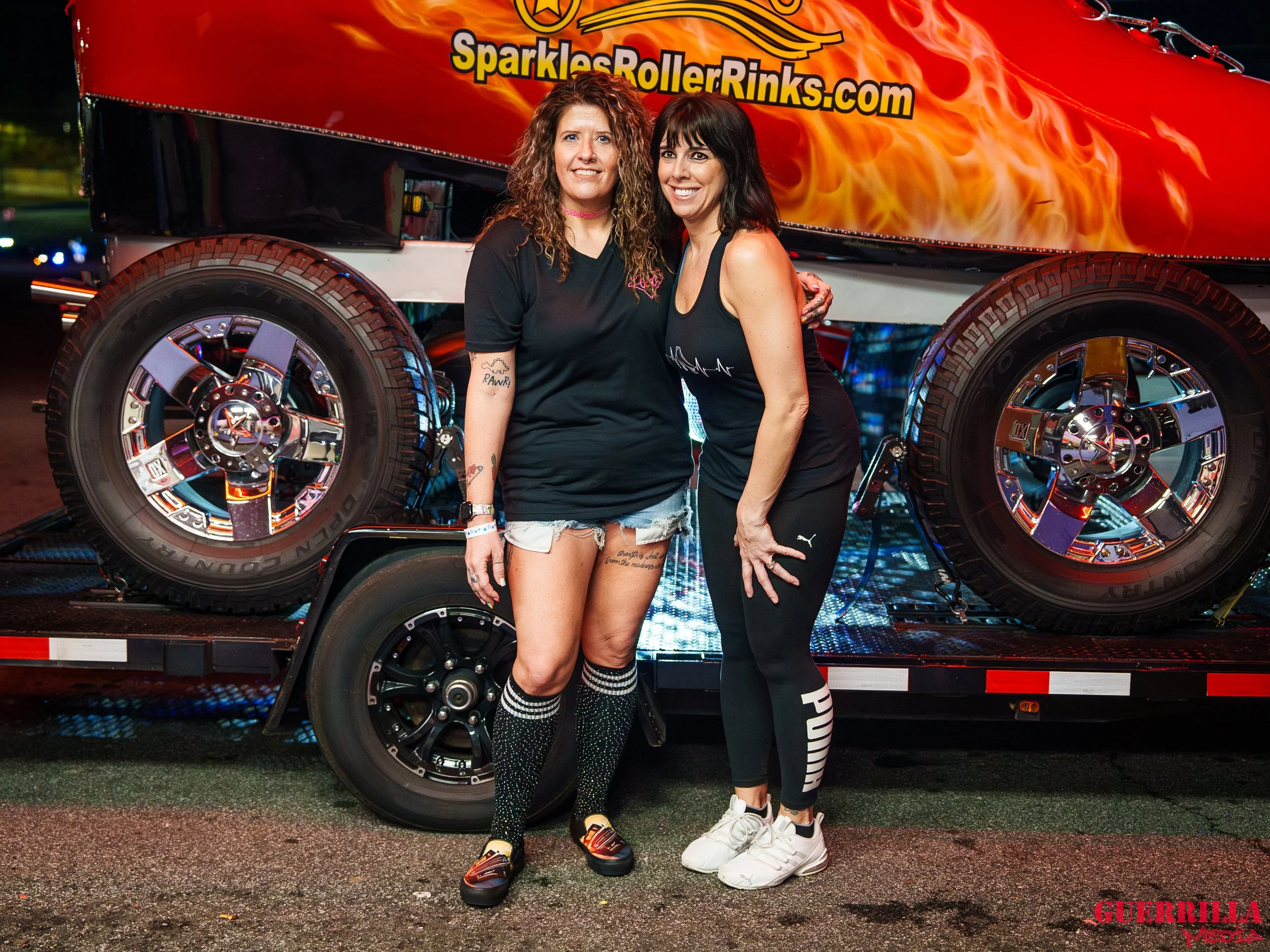 Two women standing together outdoors at night, smiling in front of a colorful roller coaster vehicle with flames and the website SparklesRollerRinks.com displayed on it.
