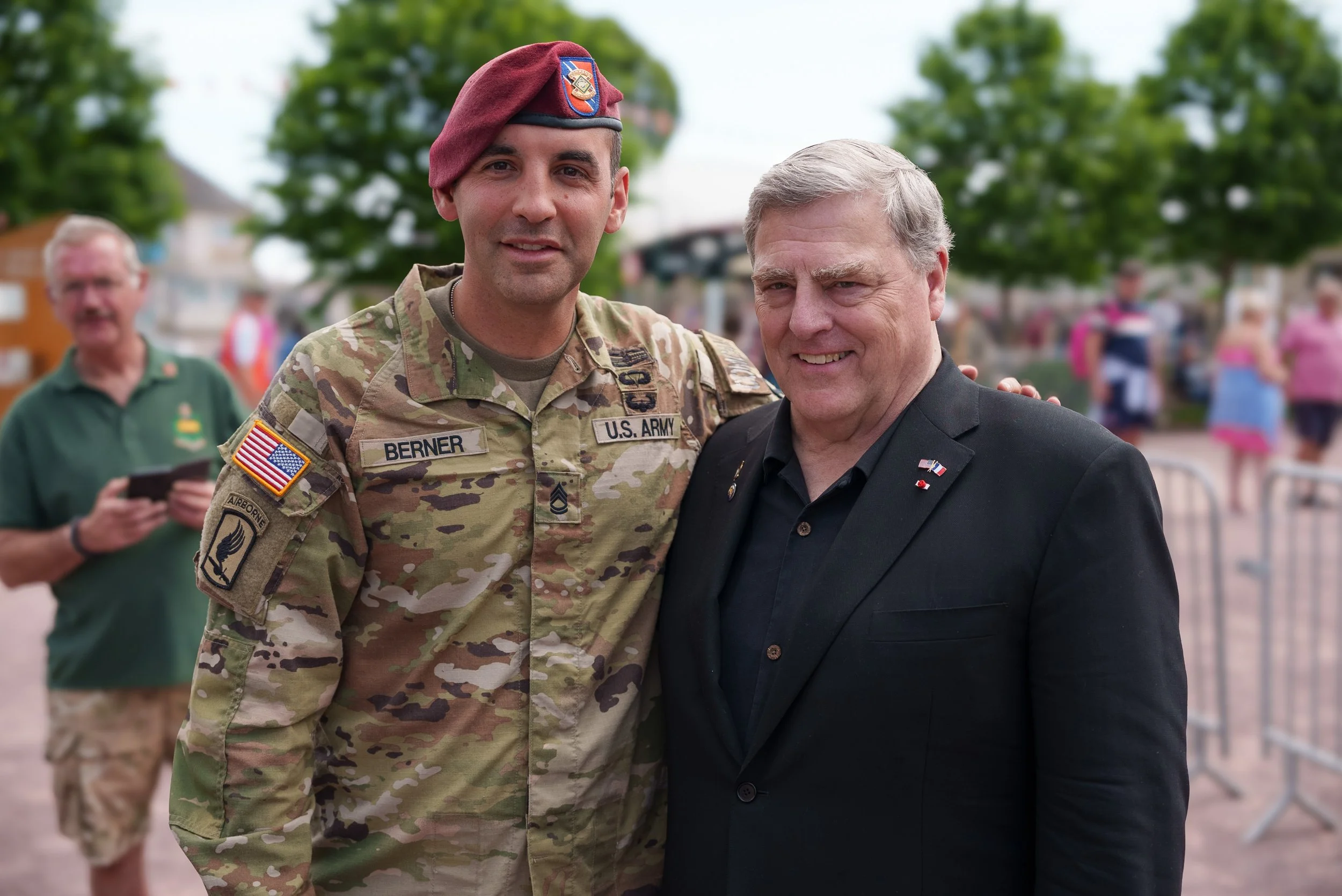 A man in U.S. Army camouflage military uniform and beret stands next to a man in a black suit outdoors during daytime. They are smiling for the photo, with a background of trees and a blurred crowd.