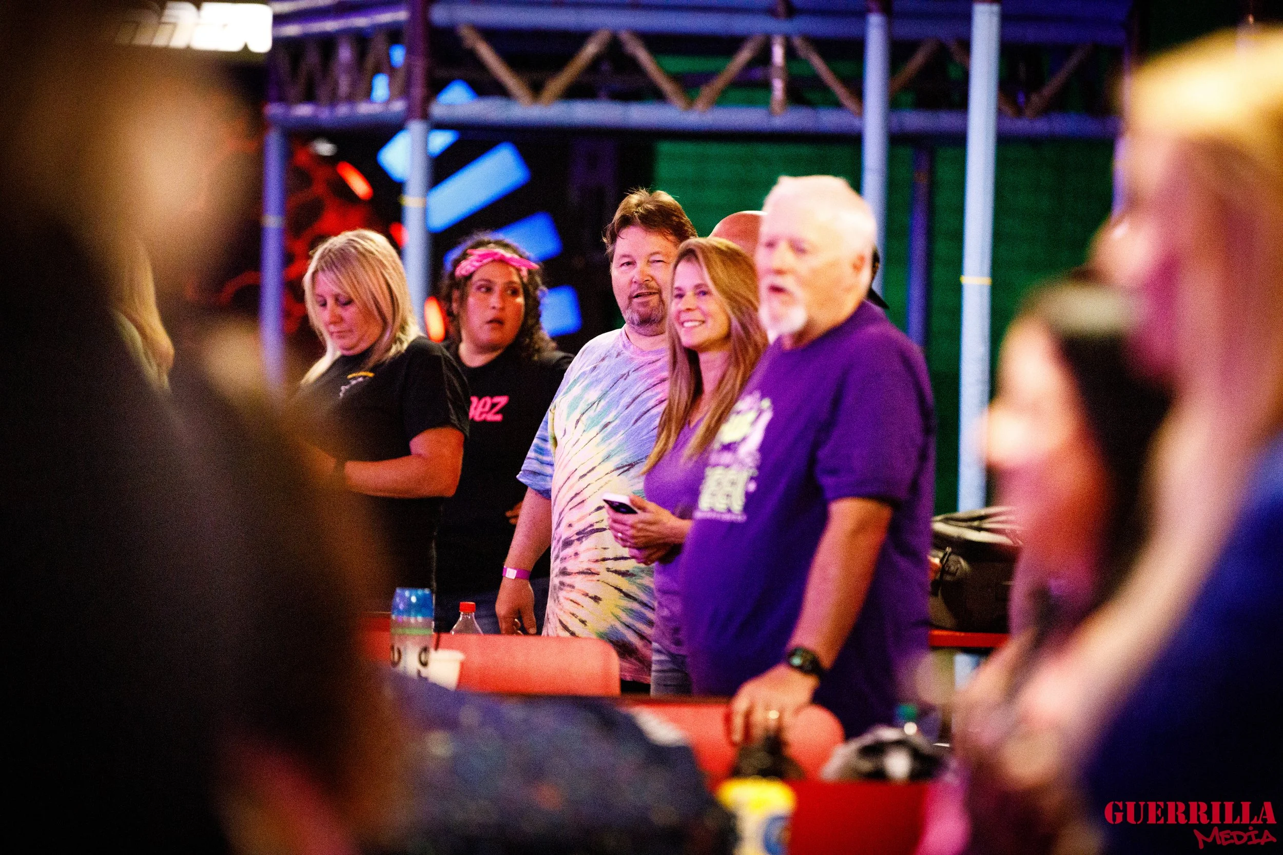 A group of people standing in a row at an indoor event, some smiling and others looking in different directions, with colorful stage lights and a metal structure in the background.
