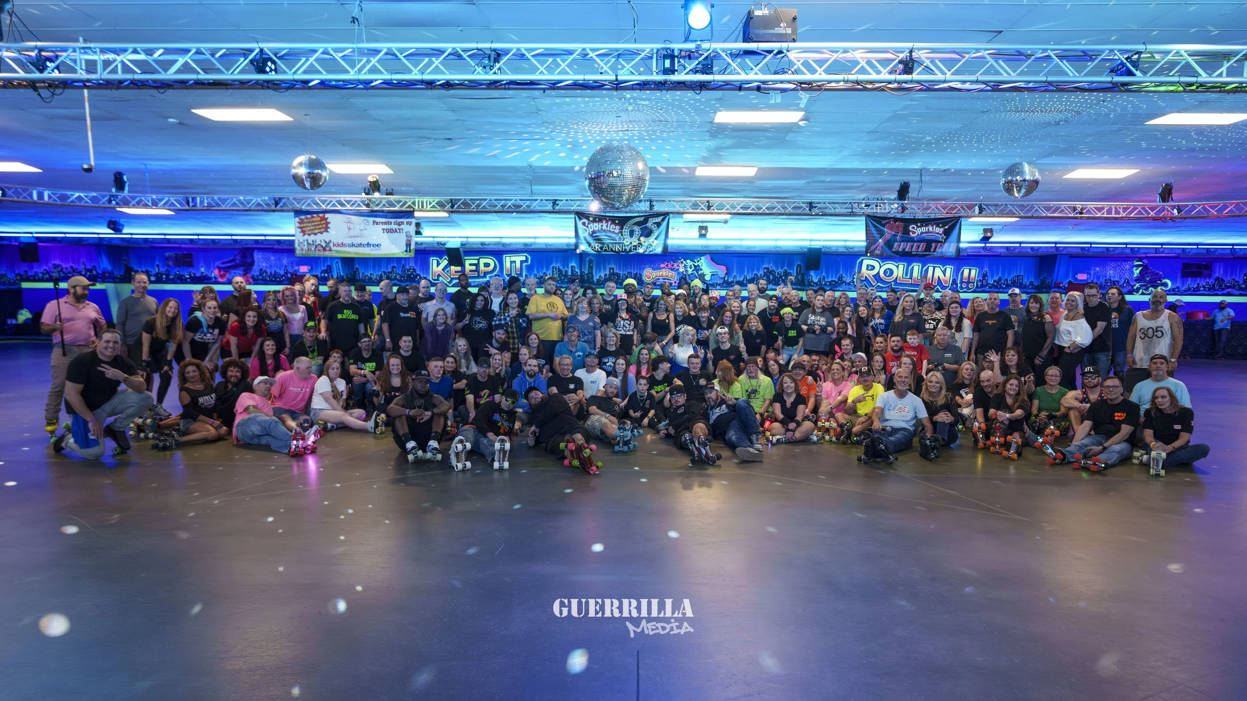 Large group of people gathered at a roller skating rink with colorful neon lighting, roller skates on some, celebrating a 6th anniversary event with a disco ball overhead and signs reading 'Keep It Rollin' and 'Sparkles Speed Team'.