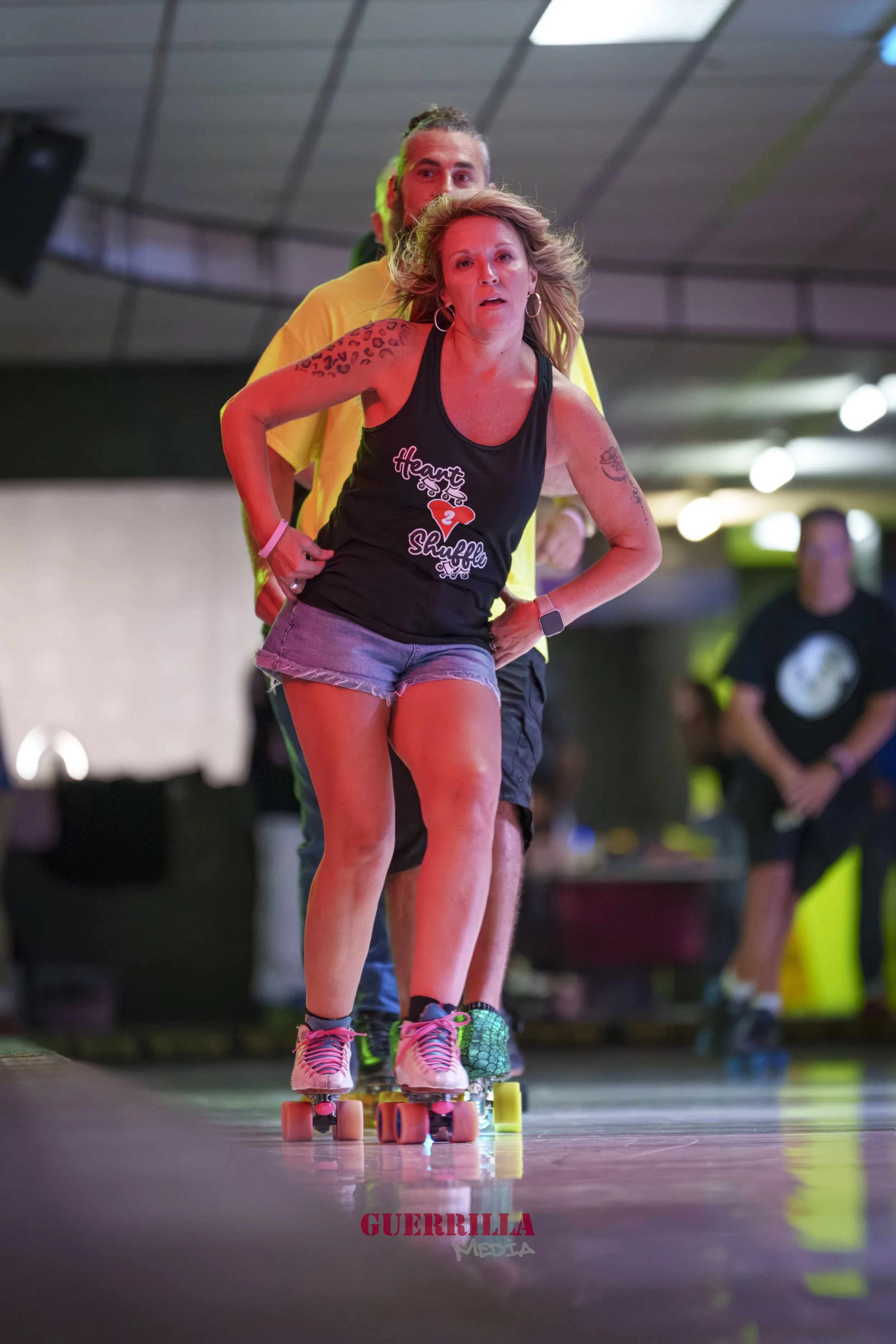 Two women roller skating indoors, one in front and one in back, with others in the background. The woman in front has blonde hair, tattoos, and is wearing a black tank top and denim shorts. The woman behind has braided hair and a yellow shirt.