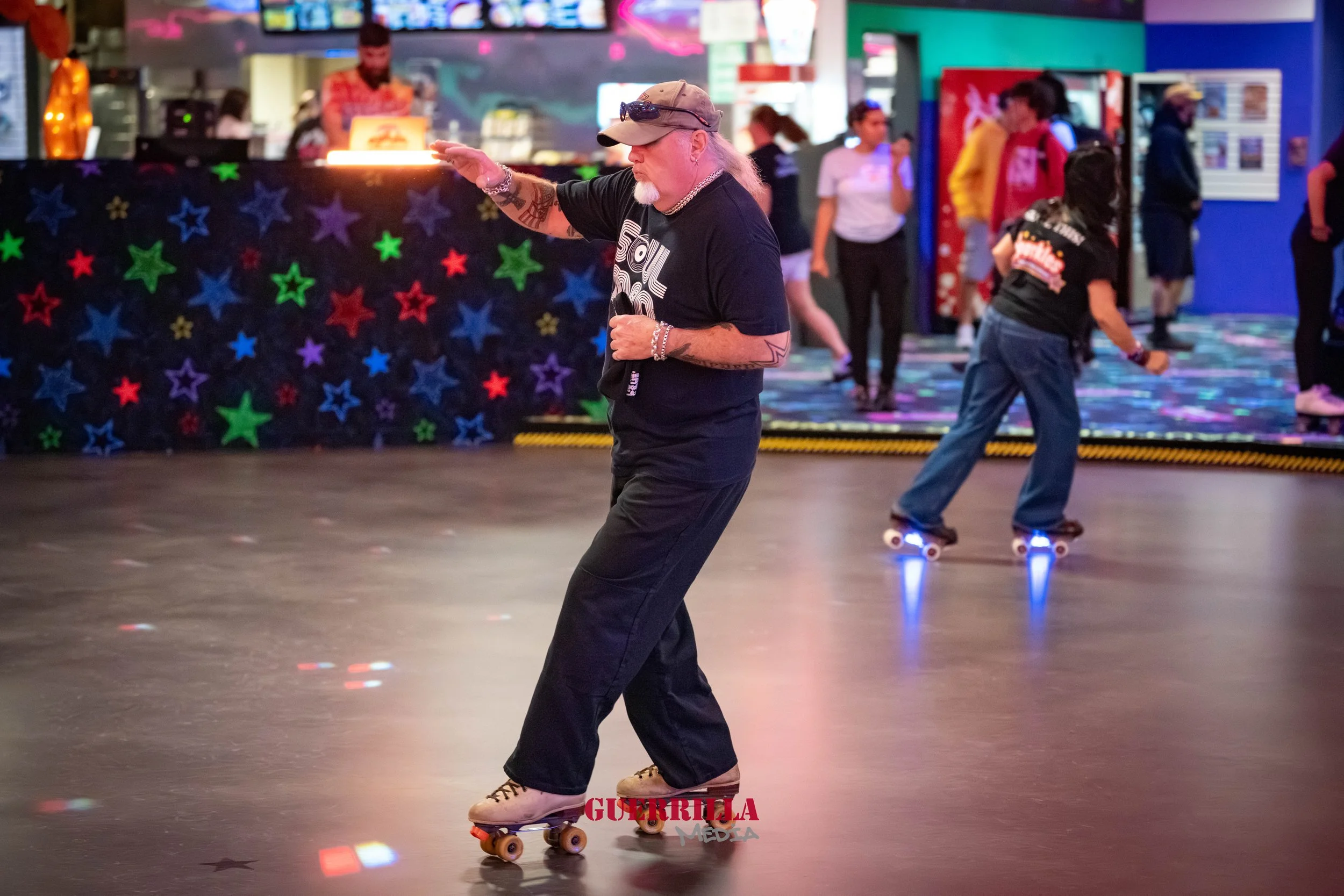 A man roller skating in an indoor rink with colorful star decorations on the wall and other skaters in the background.