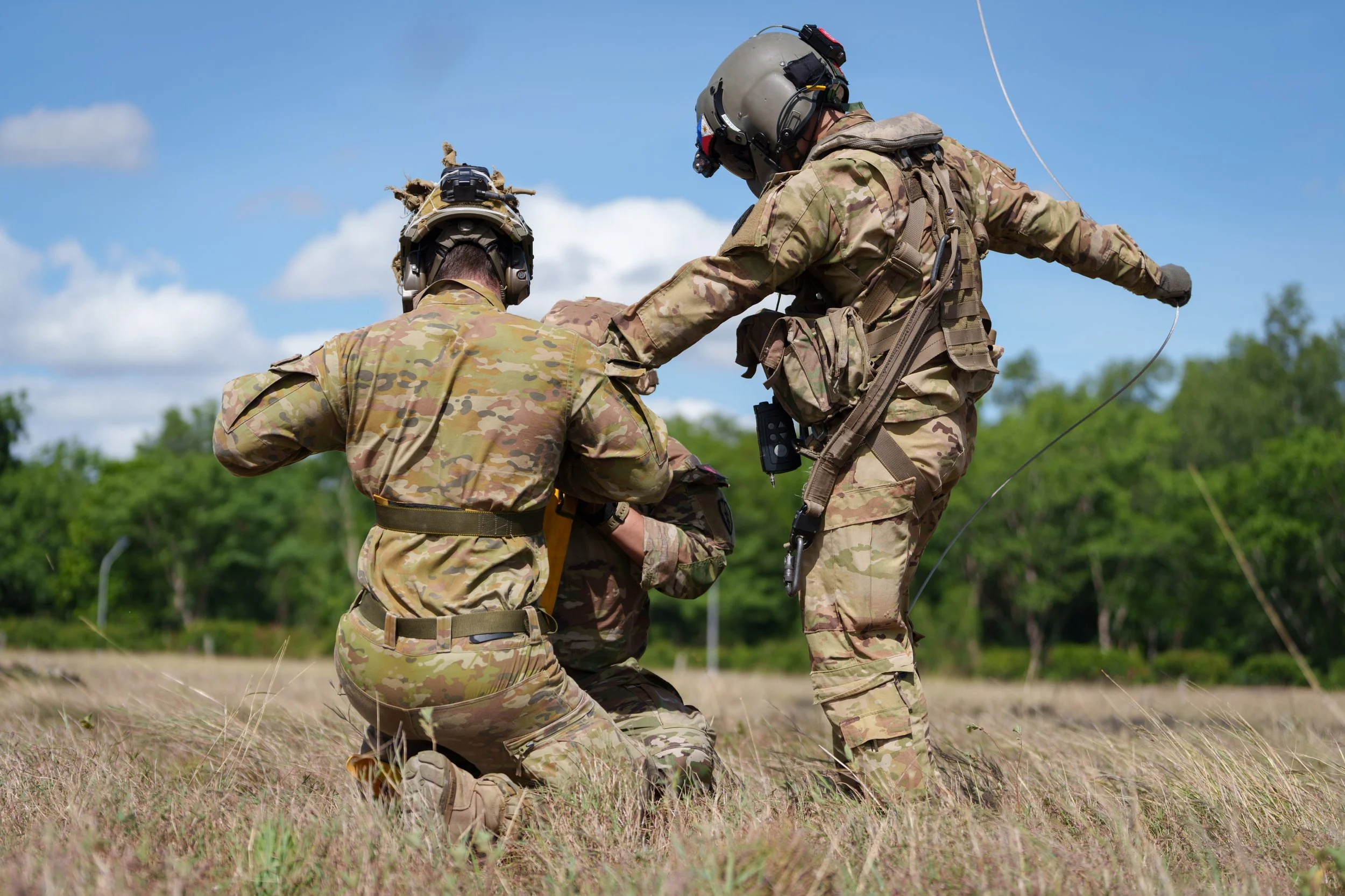 Three soldiers in camouflage uniforms participating in a battlefield exercise outdoors, with a blue sky and green trees in the background.