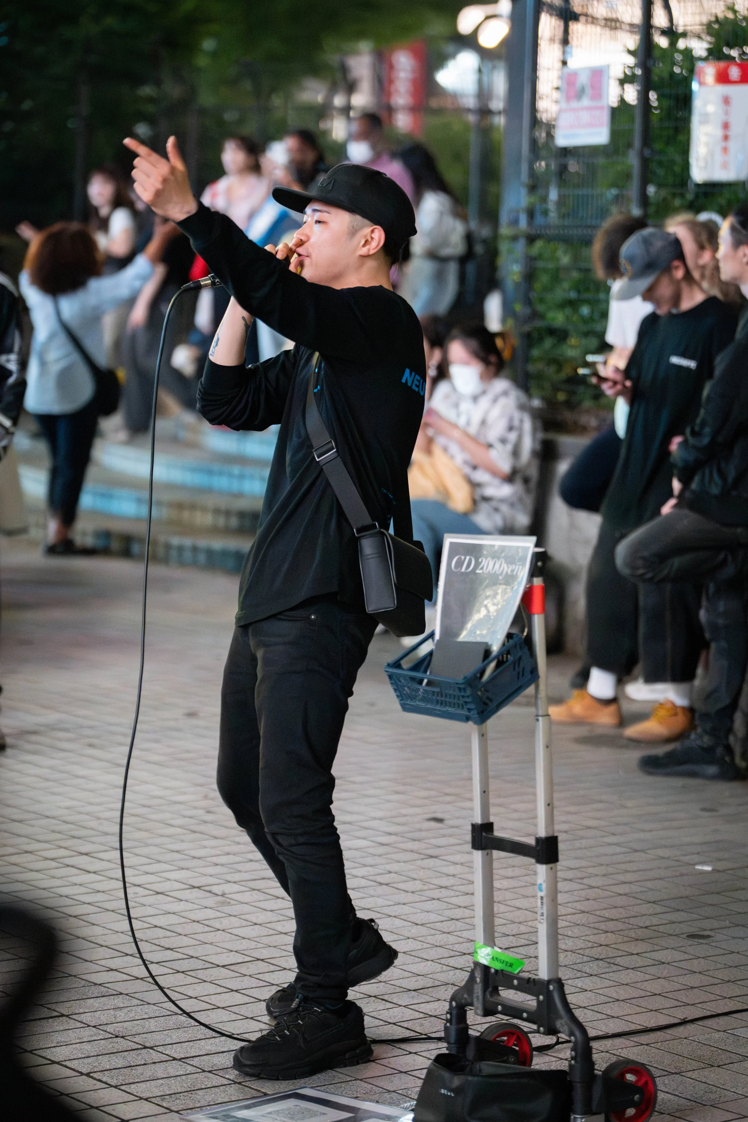 A male street performer singing into a microphone, wearing a black cap and black clothing, with a crowd of people around him, some seated and some standing, all wearing masks.