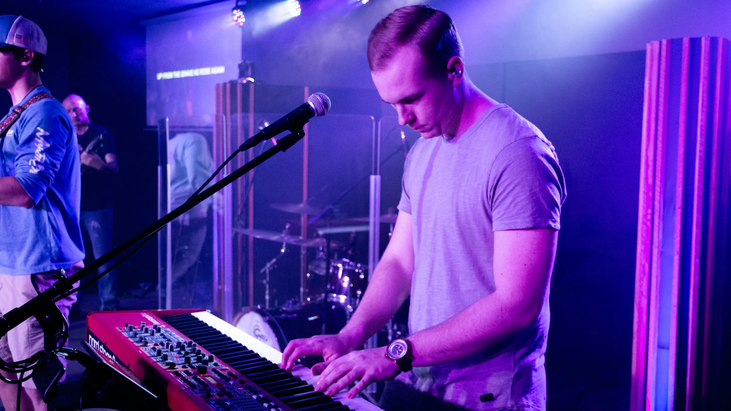 Young man playing a keyboard on stage with purple and blue lighting, microphone in front, other band members in background.