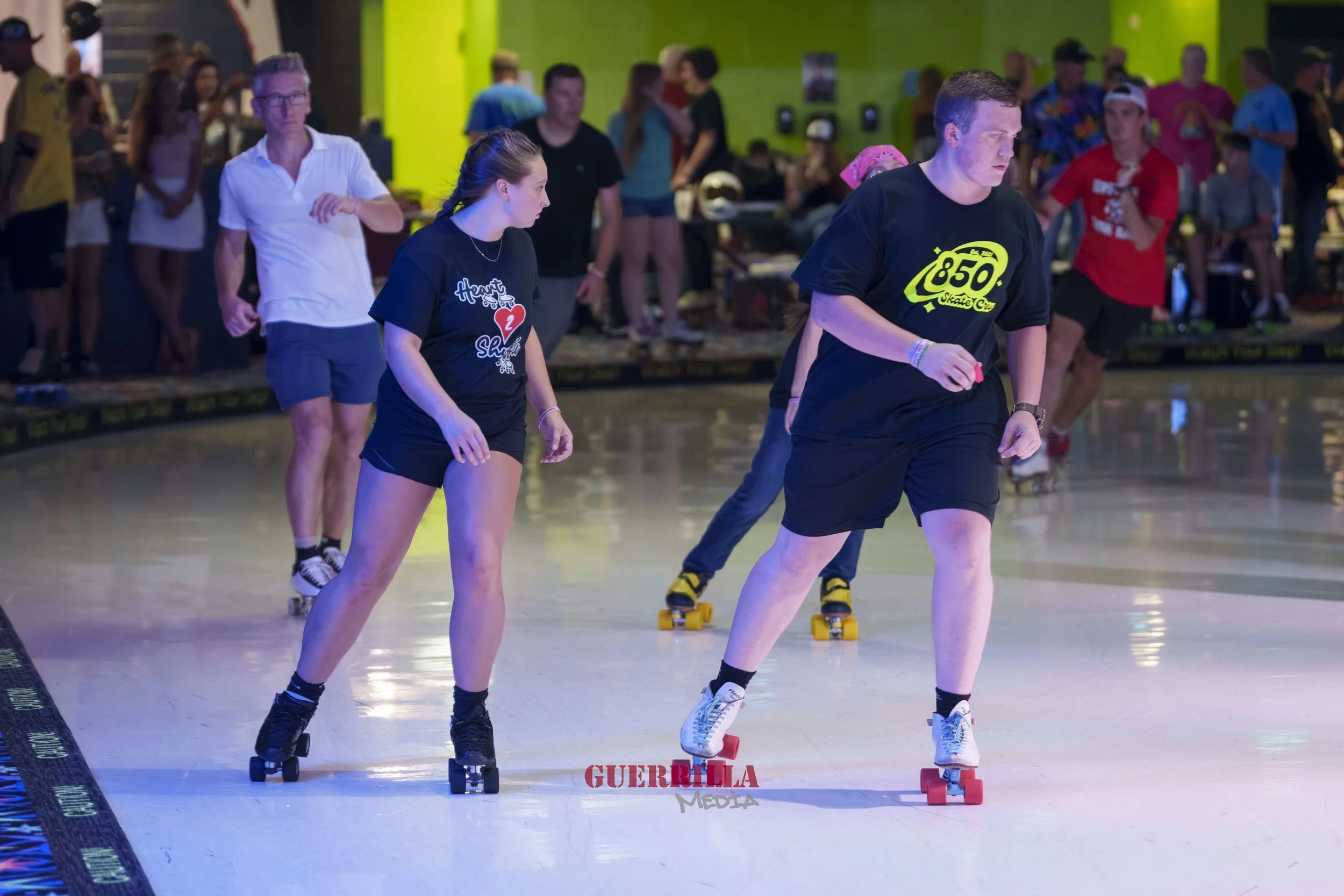 People roller skating indoors, with one person wearing a black shirt with the number 850 and another in a black shirt with a heart and heart logo, in a skate rink with spectators in the background.