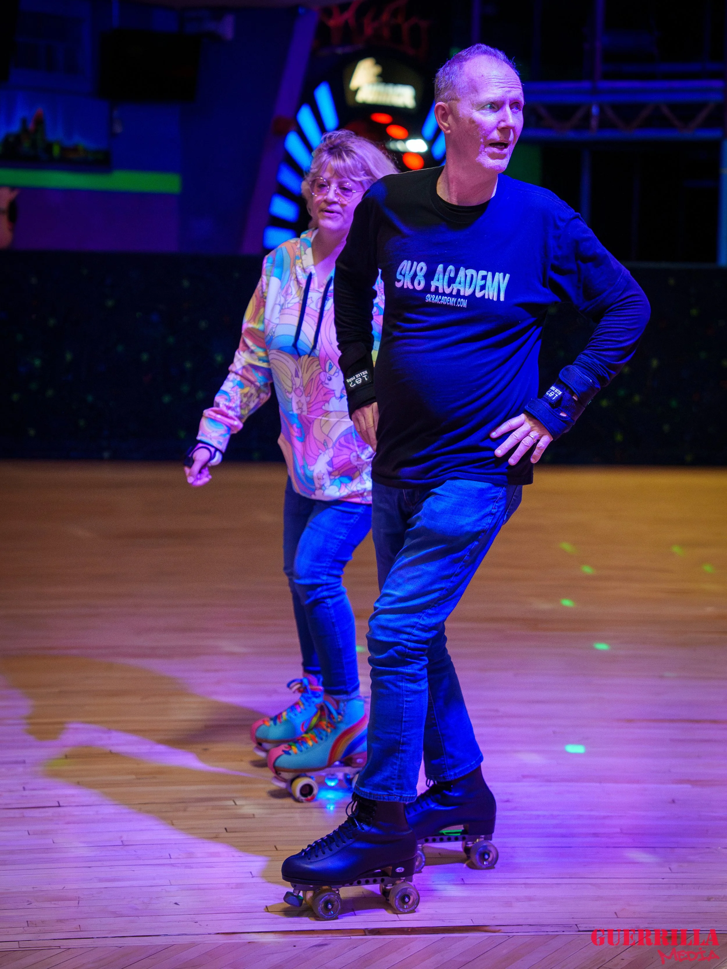 Two people roller skating indoors under colorful neon lighting, with a man in a black shirt and a woman in a colorful hoodie.