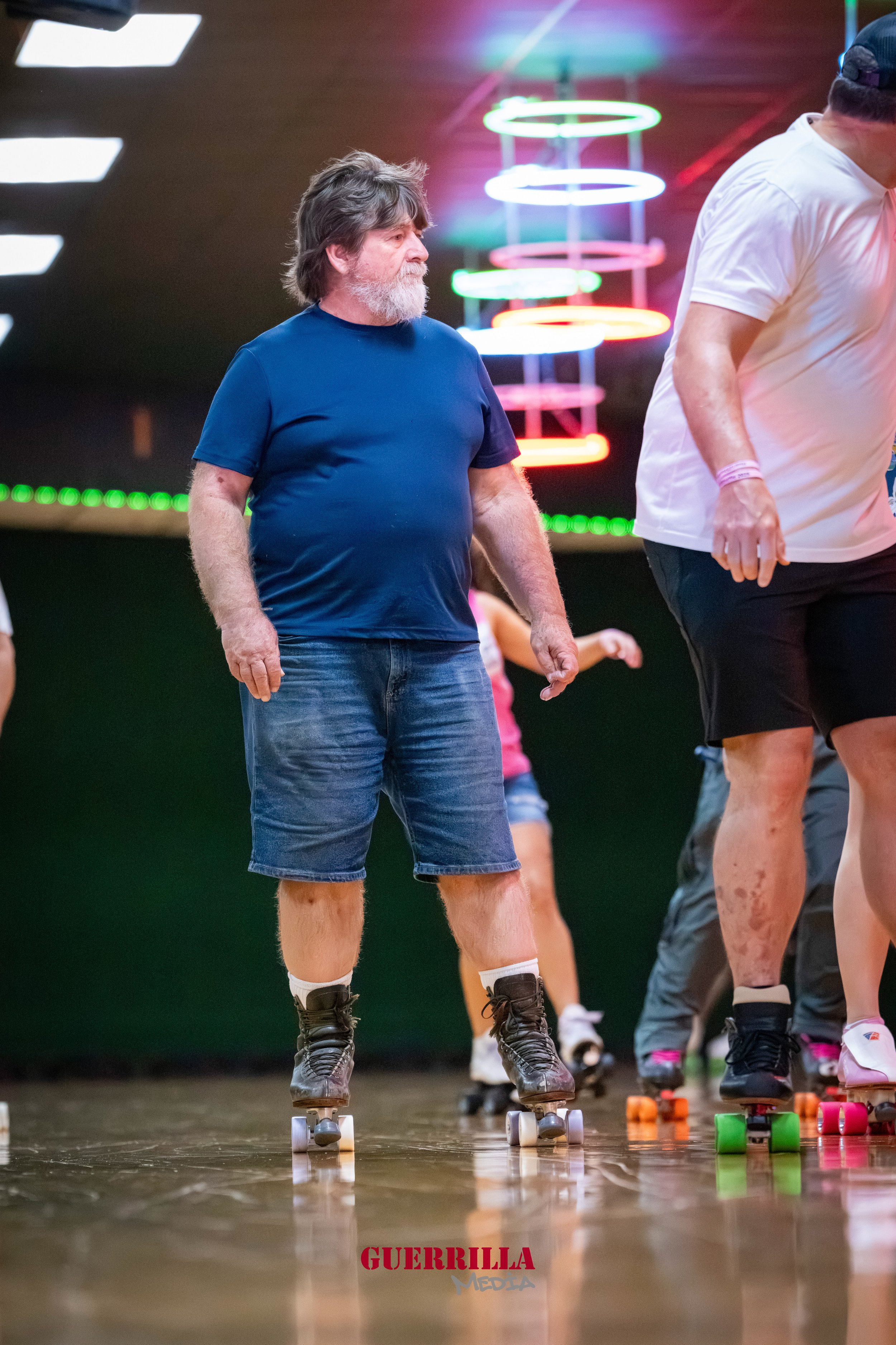 A man with long hair, beard, wearing a blue t-shirt, denim shorts, and roller skates, skating indoors at a neon-lit roller rink.
