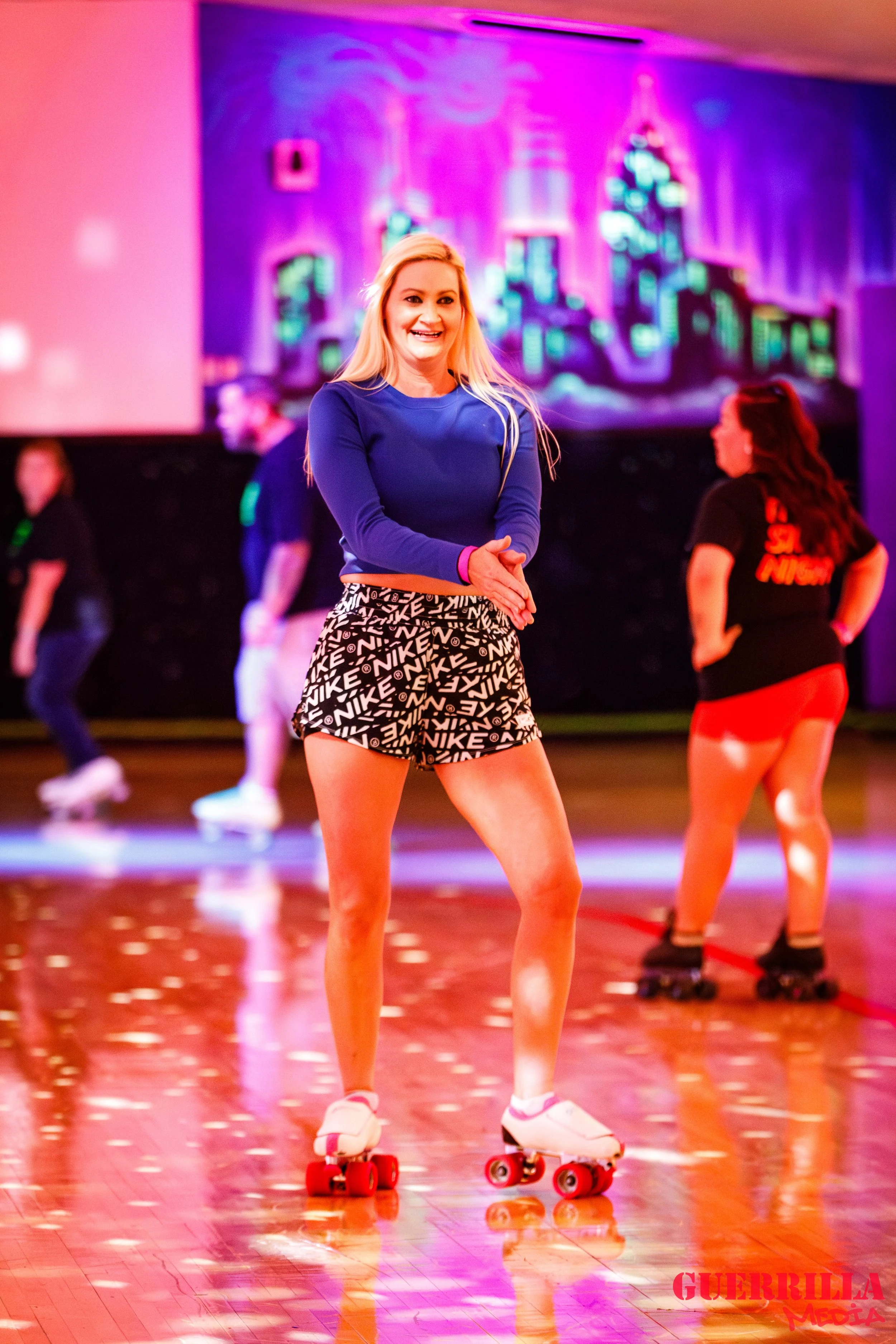 A young woman roller skating inside a roller disco rink, smiling, wearing a blue long-sleeve top, Nike patterned shorts, and white roller skates, with a colorful cityscape mural in purple and blue hues on the wall behind her.