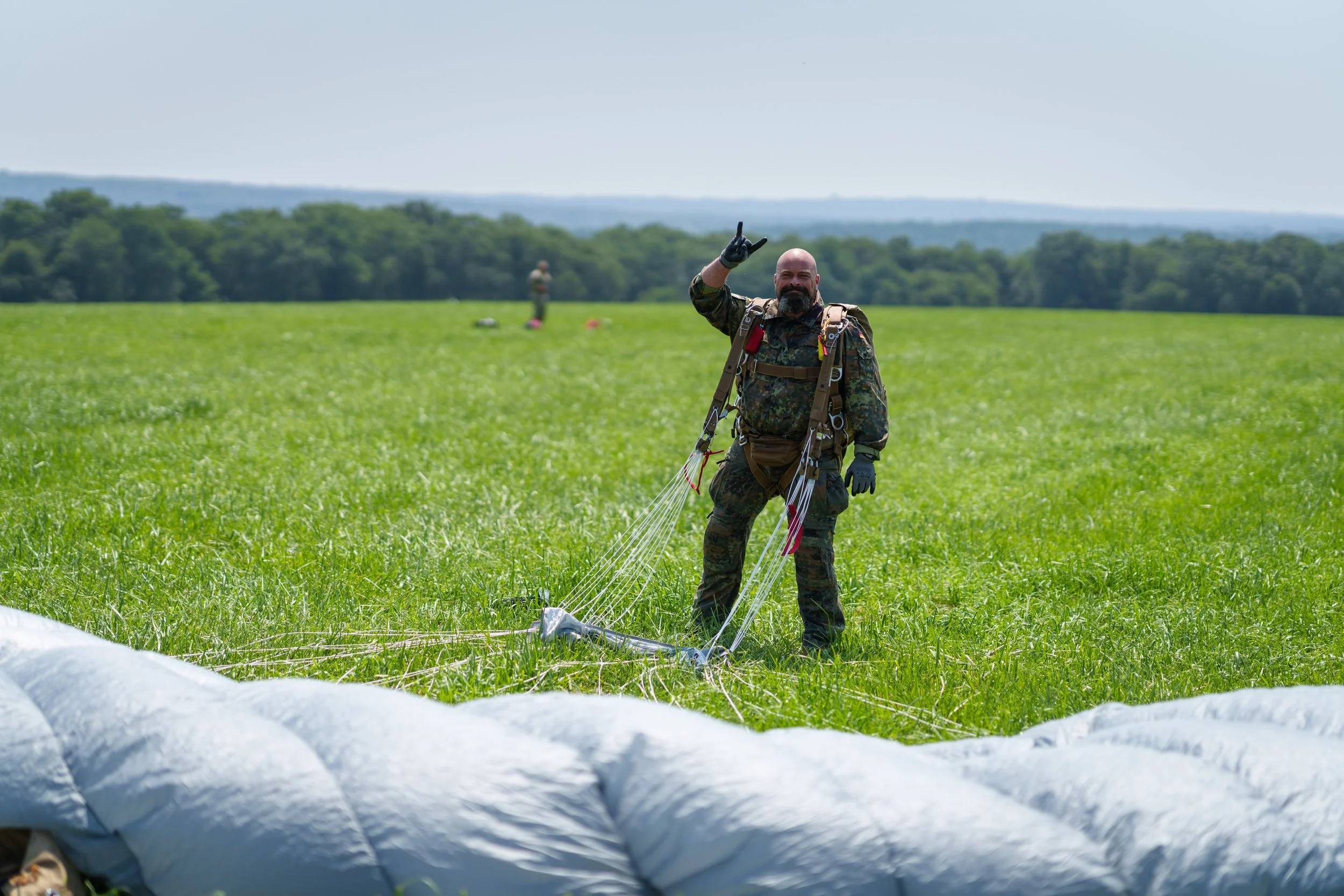 A military parachutist standing in a grassy field, giving a peace sign, with descending parachute in the foreground and a clear sky in the background.