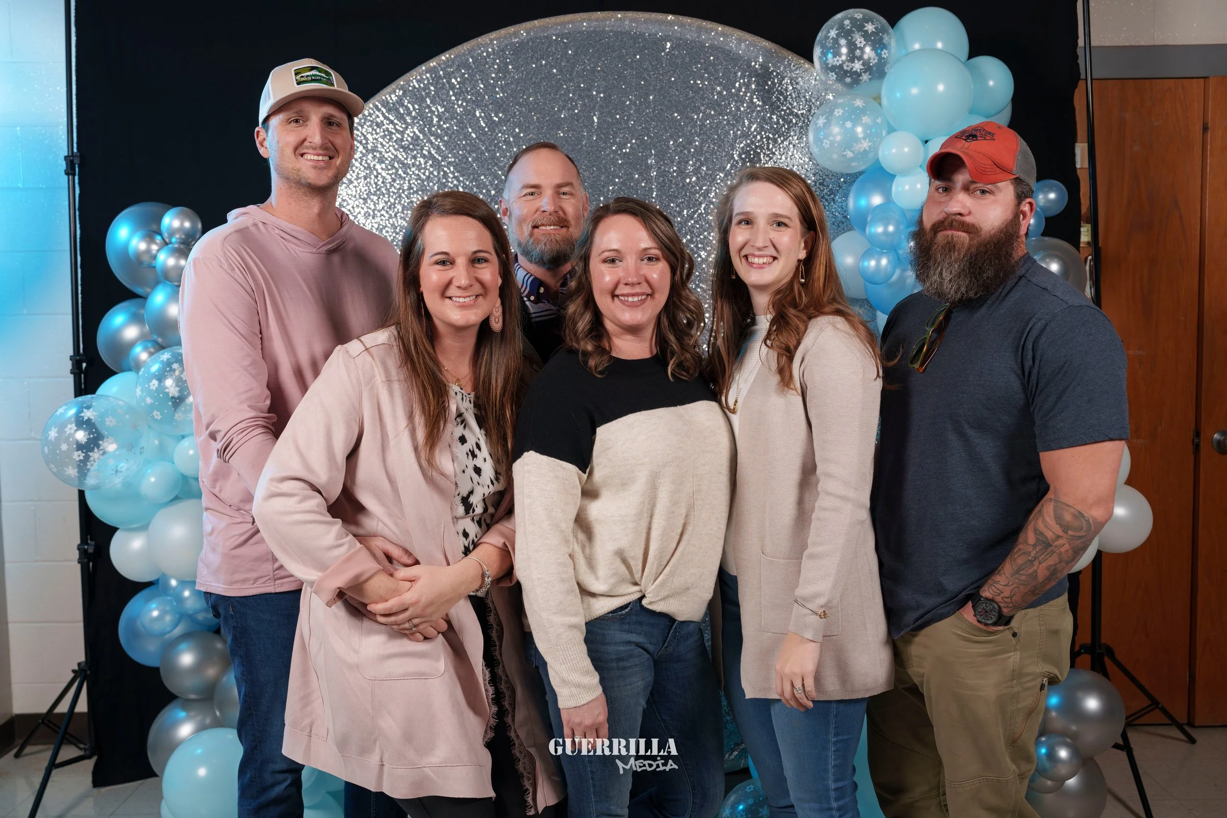 Group of six people smiling, standing in front of a silver glittery backdrop decorated with blue and silver balloons for a celebration.