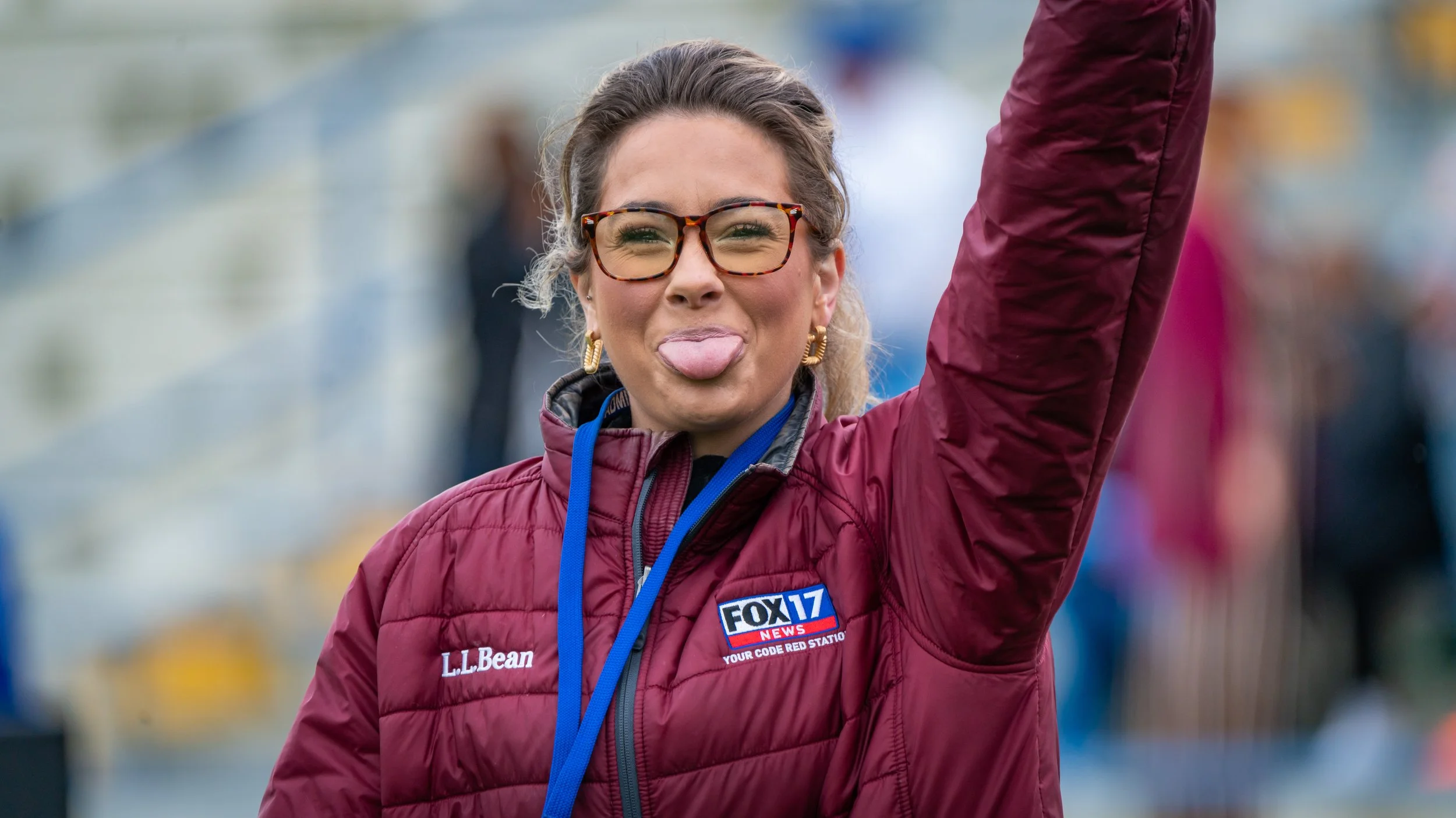 A woman with glasses and earrings sticking out her tongue and raising her arm at an outdoor event. She is wearing a maroon jacket with a FOX 17 News patch and a blue lanyard.