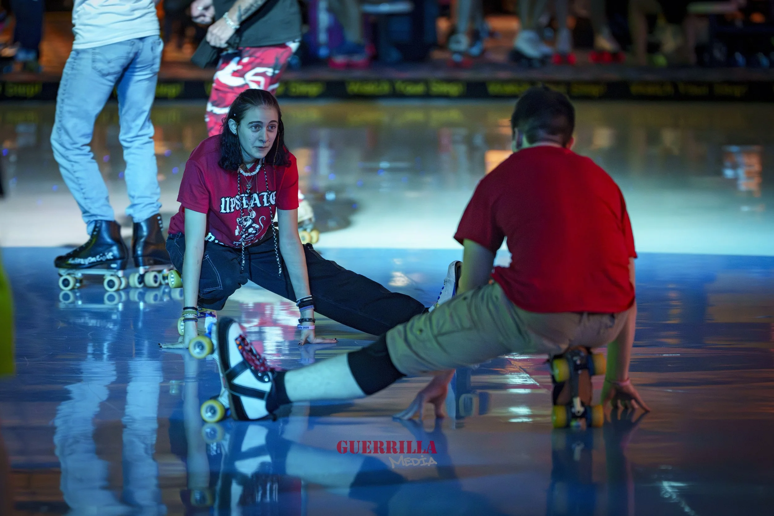 A young woman and a young man in roller skates are on the floor of an indoor roller skating rink, sitting with their legs extended and leaning on their hands, while other skaters are visible in the background. The woman has dark, shoulder-length hair