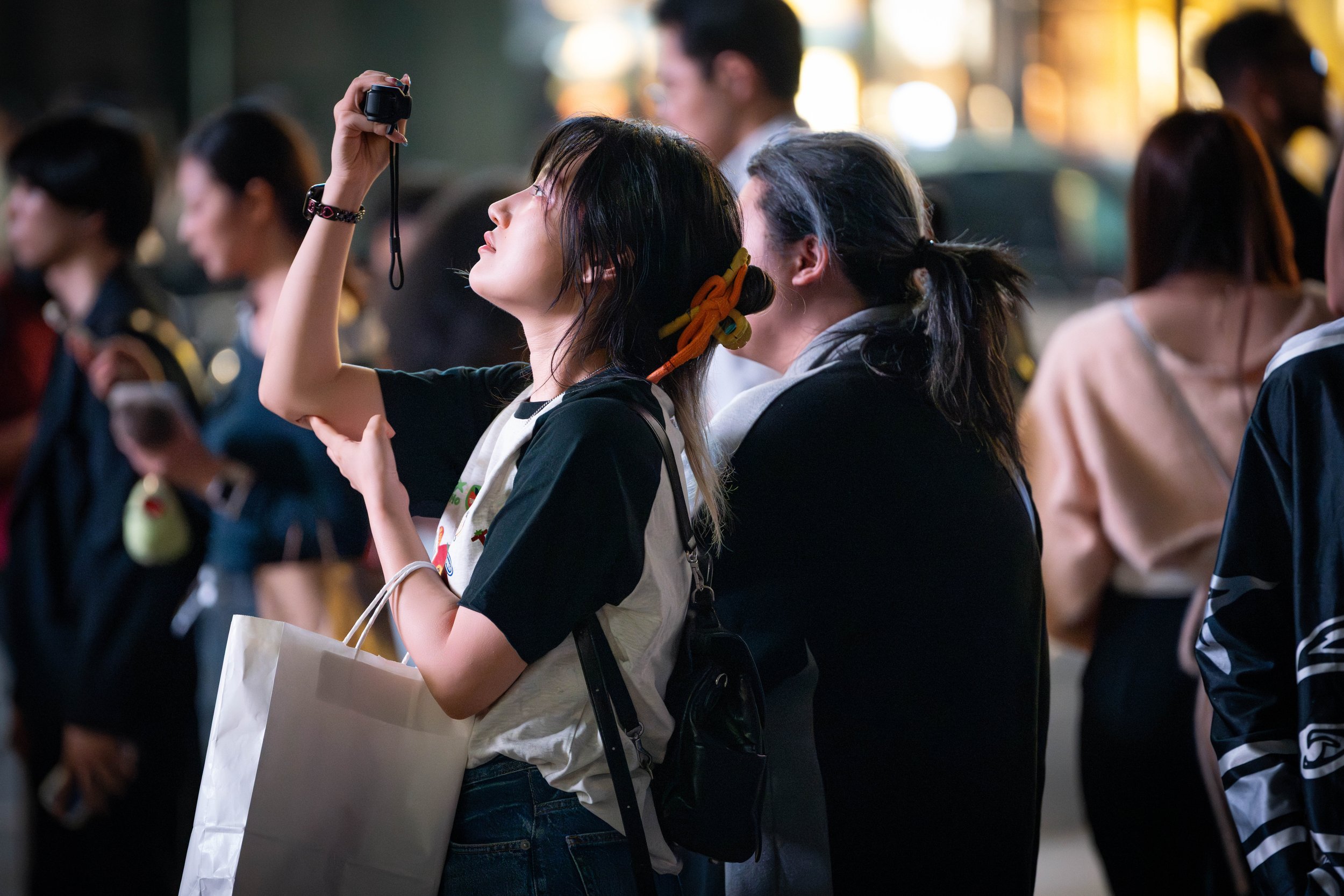 A young woman with dark hair taking a photo with a camera in a crowd during nighttime.